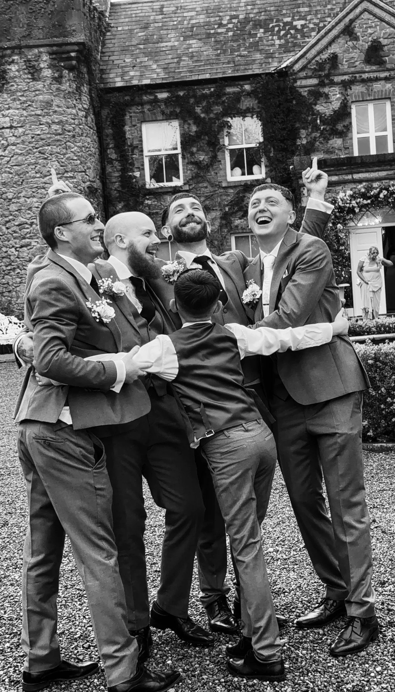 Group of men in suits celebrating outdoors at a wedding, smiling and hugging with a historic stone building in the background.