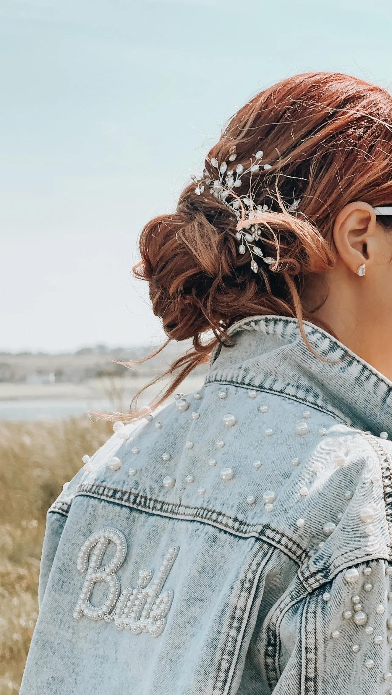 A woman with styled red hair wearing a denim jacket adorned with pearls, eyeshadow, and pearl earrings, standing outdoors in a field.