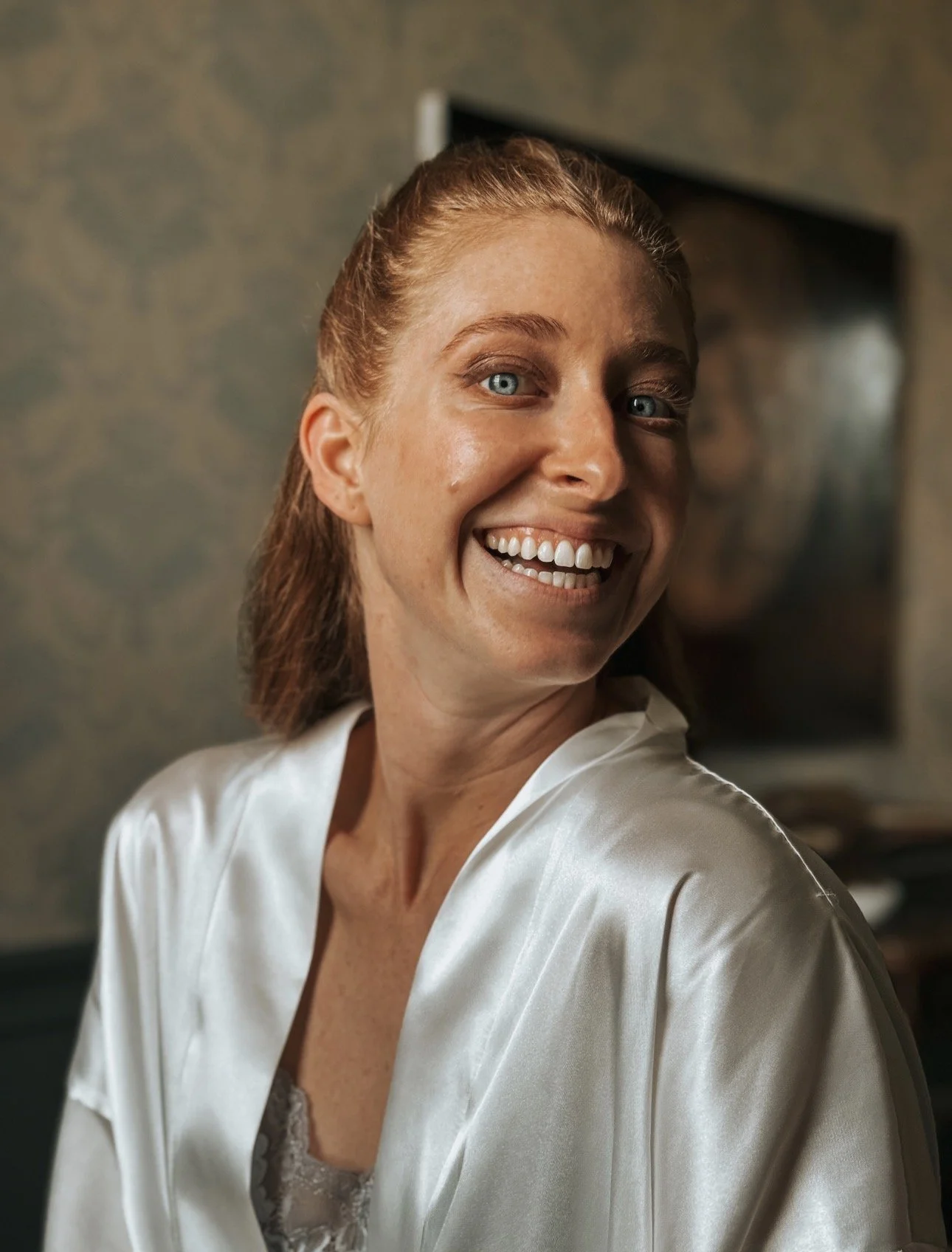 A smiling woman with red hair and blue eyes wearing a white satin robe getting ready for her wedding morning in Carton House, Maynooth. Wedding Content Creator Ireland - Grey Acre Studios