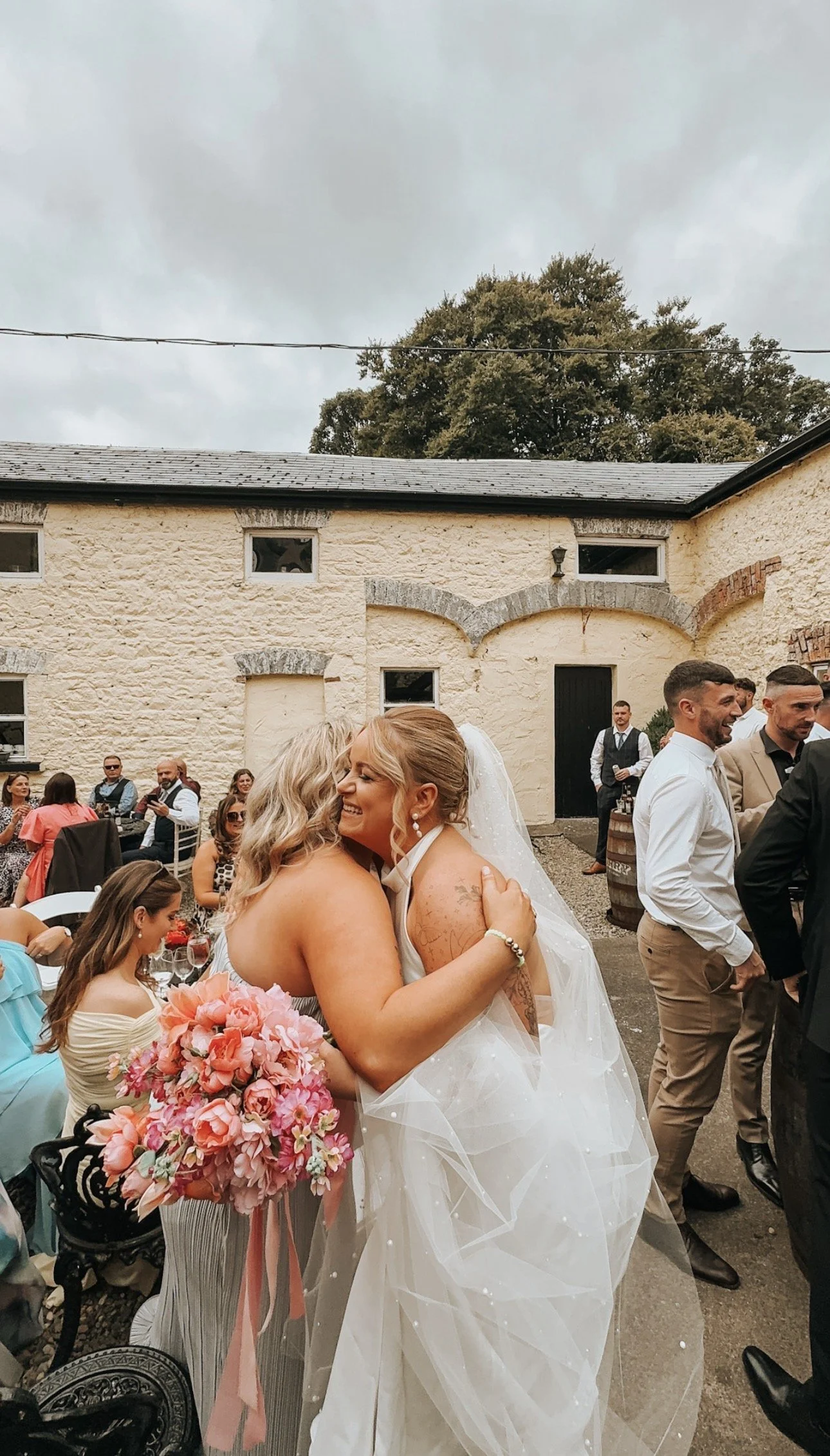 Two women hugging at a wedding reception outdoors, with guests seated at tables in the background. Captured by Wedding Content Creator Grey Acre Studios, in County Louth, Ireland 