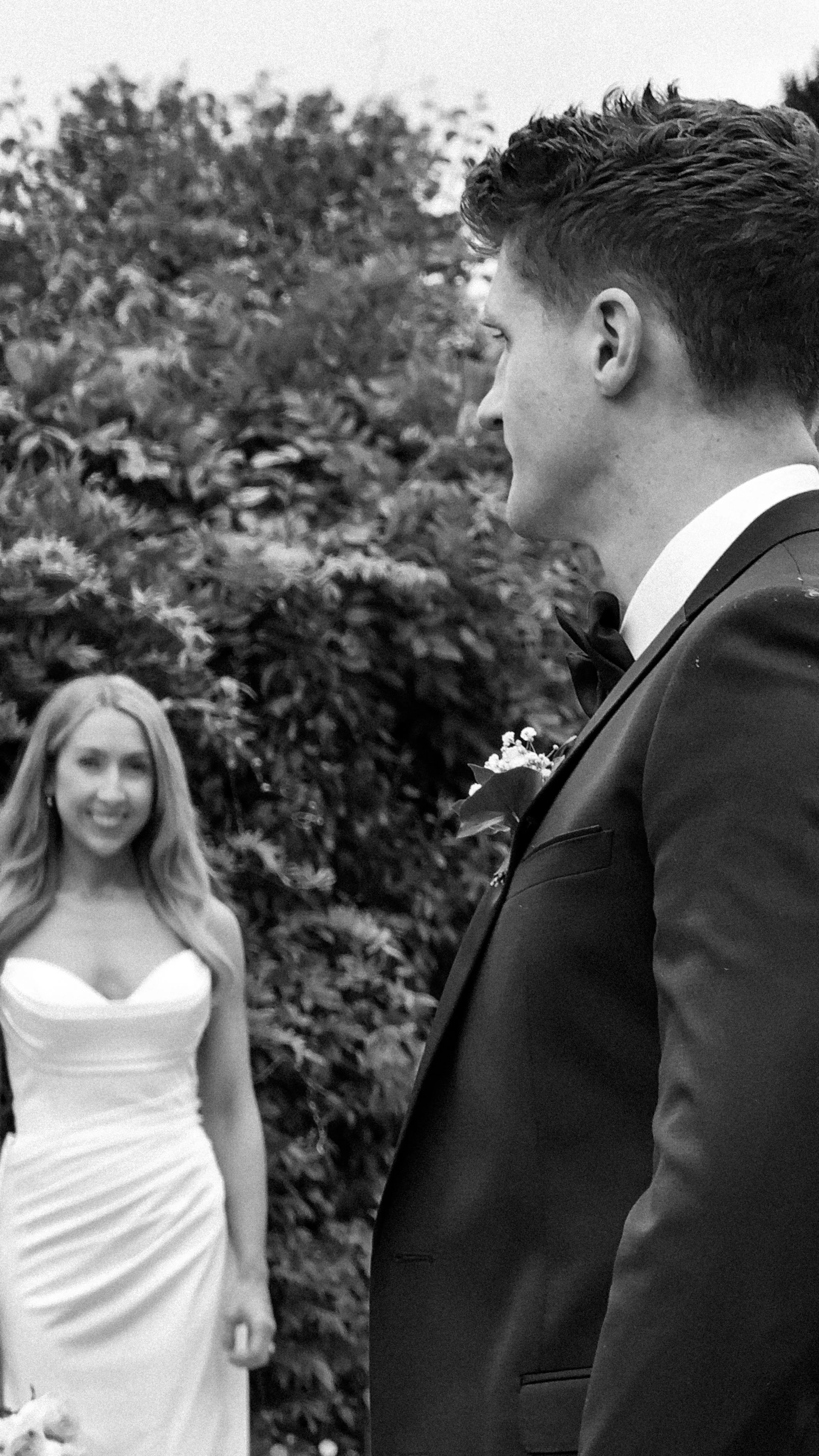 A black and white photo of a bride and groom. The bride is smiling in the background, wearing a strapless white wedding dress, and the groom is in the foreground, dressed in a black tuxedo with a bow tie and boutonniere at Kilkea Castle, Kildare