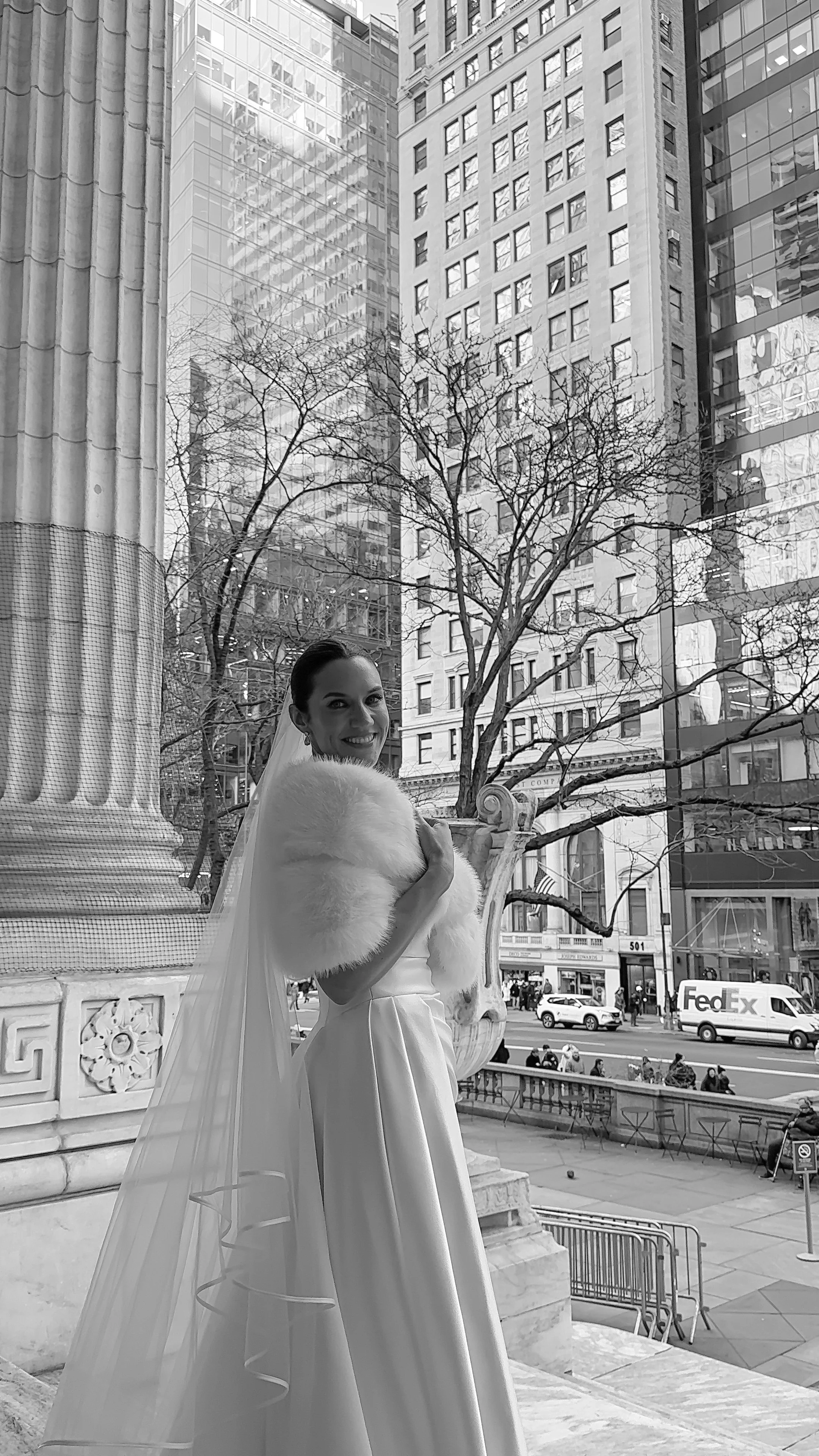 A woman in a wedding dress and fur stole standing outside in front of a city building, smiling at the camera.