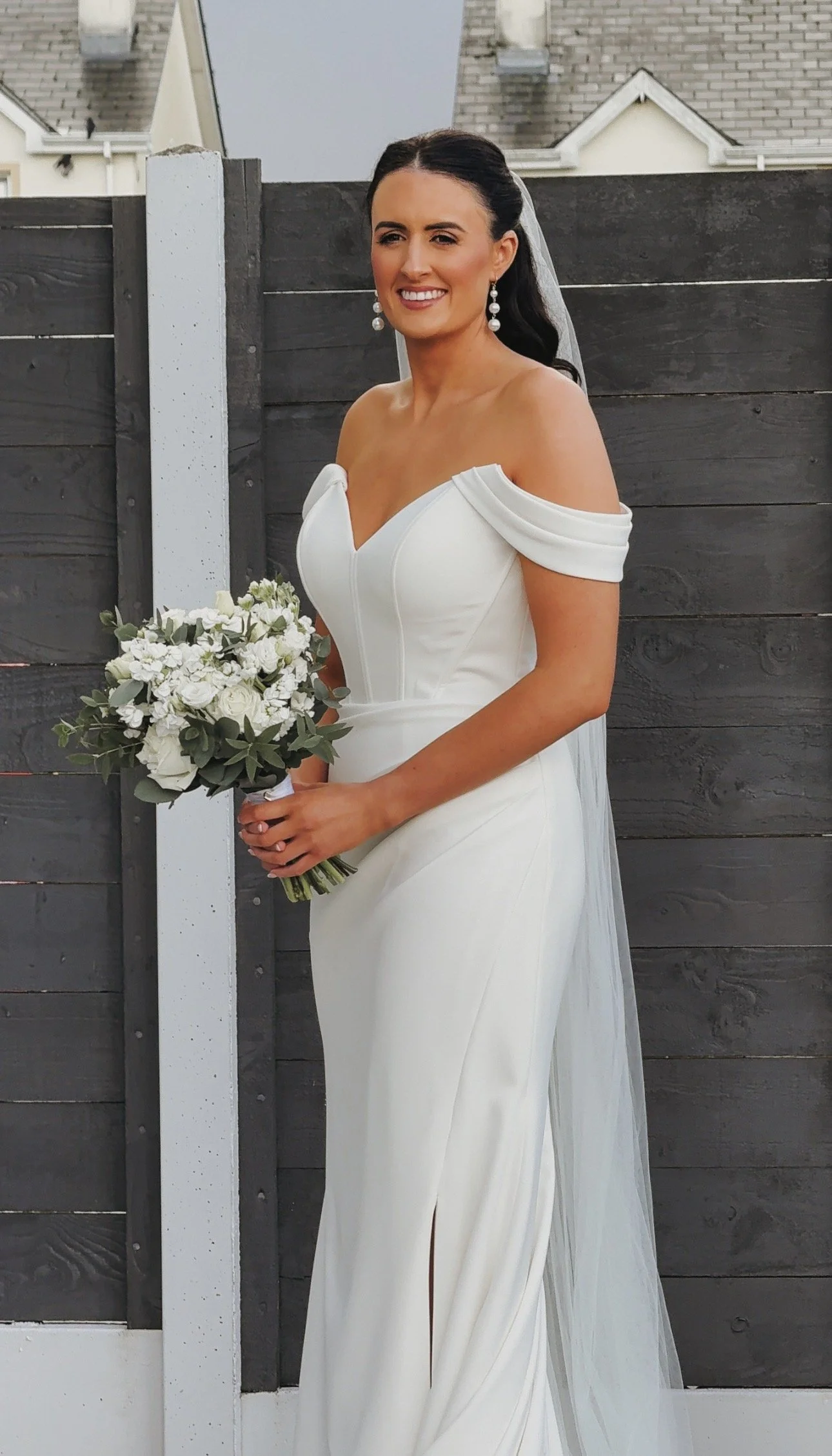 A bride in an off-shoulder white wedding gown holding a bouquet of white flowers and greenery, standing outdoors in front of a black wooden fence in Co. Sligo. Wedding Content Creator Ireland.