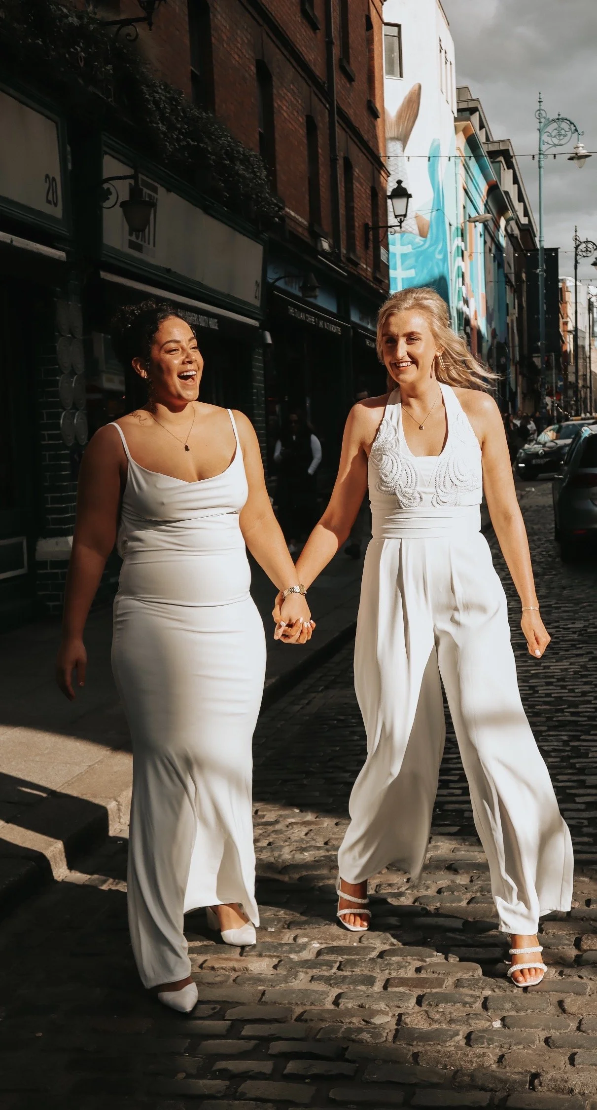 Dublin City Wedding. Two women dressed in white, holding hands, walking on a cobblestone street, smiling and enjoying each other's company with urban buildings and a mural in the background. Wedding Content.