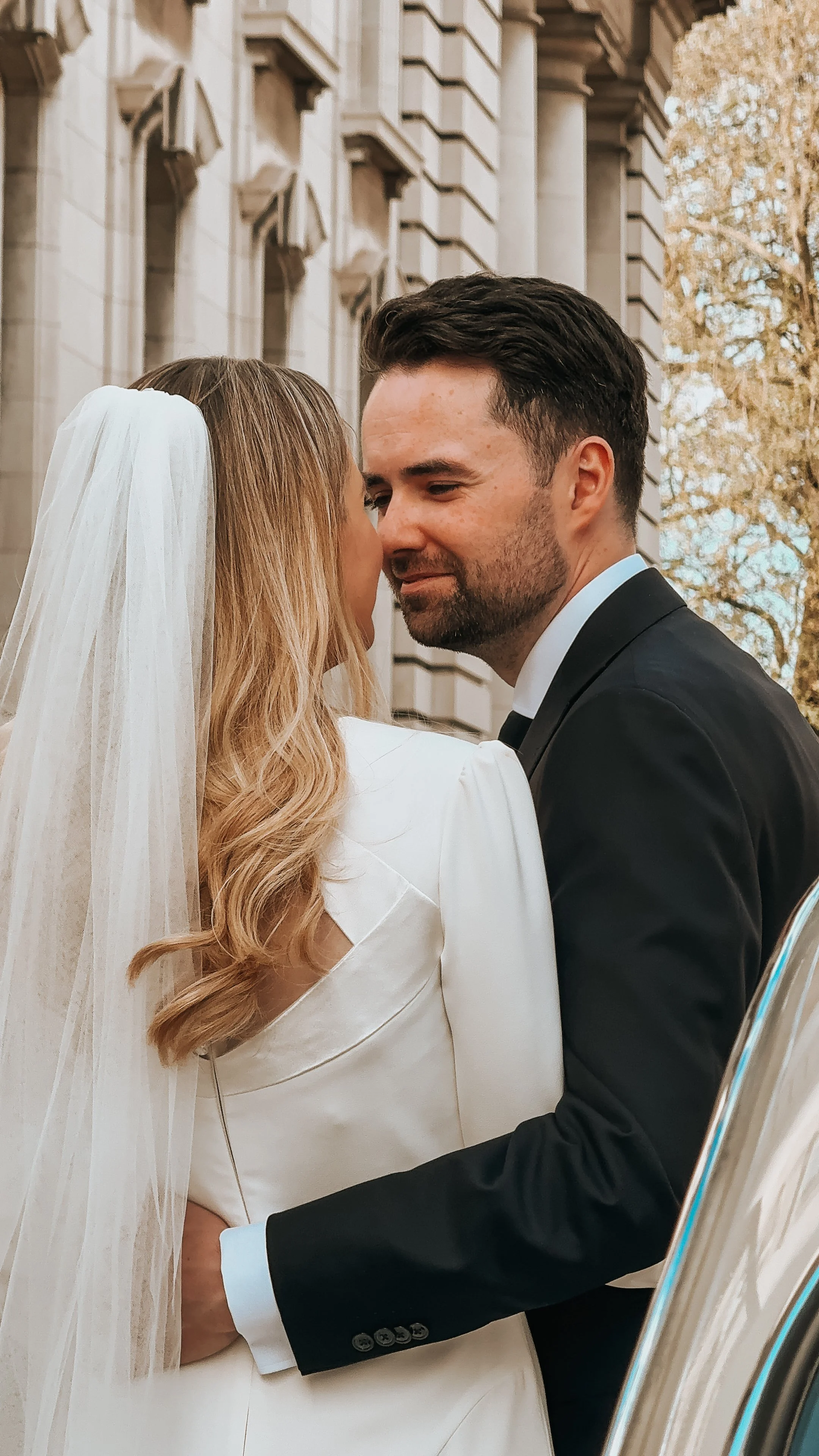 A bride and groom are close together, sharing an intimate moment during their wedding outside a historic building with detailed architecture and autumn trees in the background. Dublin City Wedding, Wedding Content Creator Ireland 