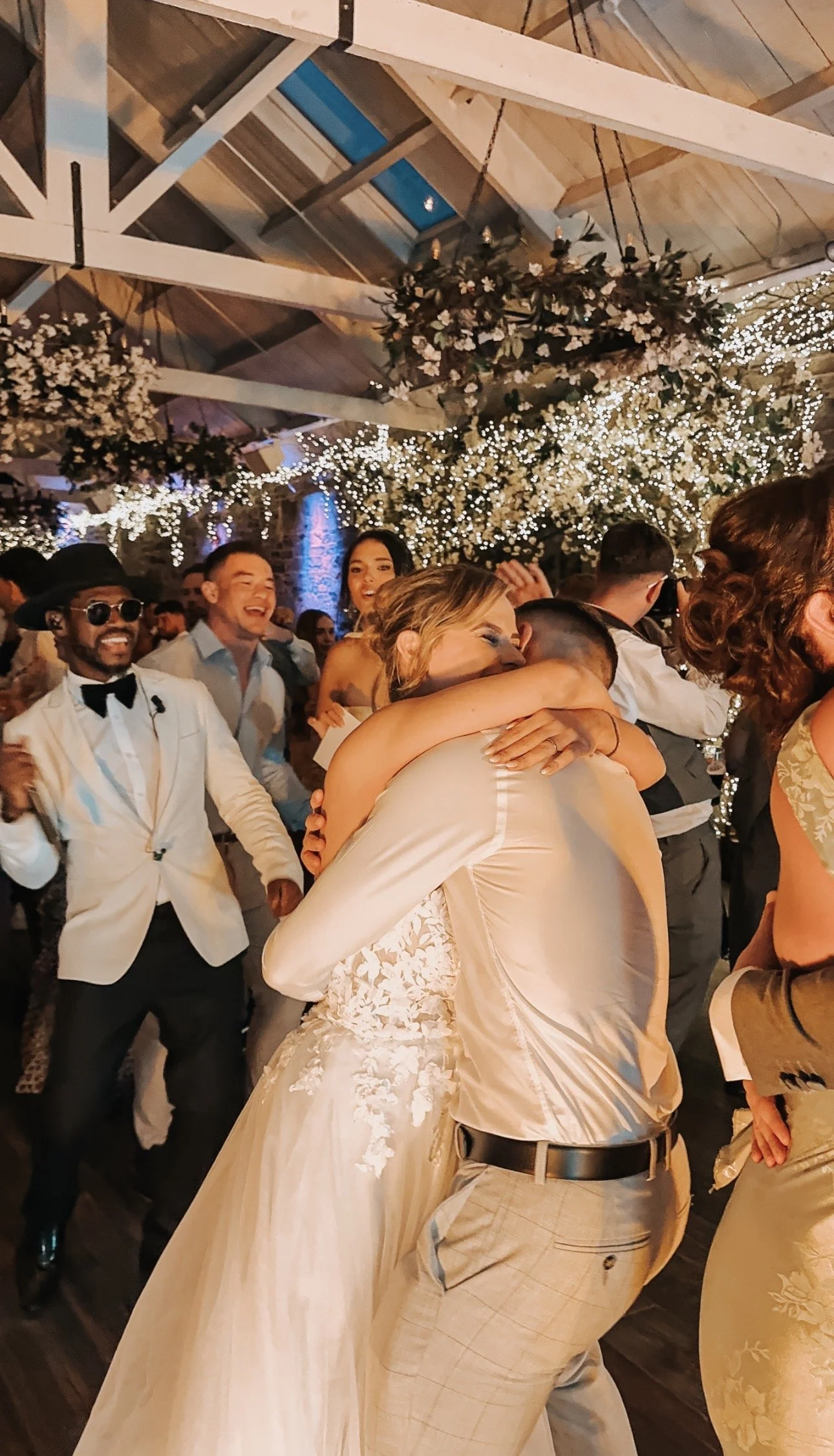 Couple embracing on a dance floor at a wedding reception, surrounded by smiling guests, with twinkling string lights and floral decorations overhead.