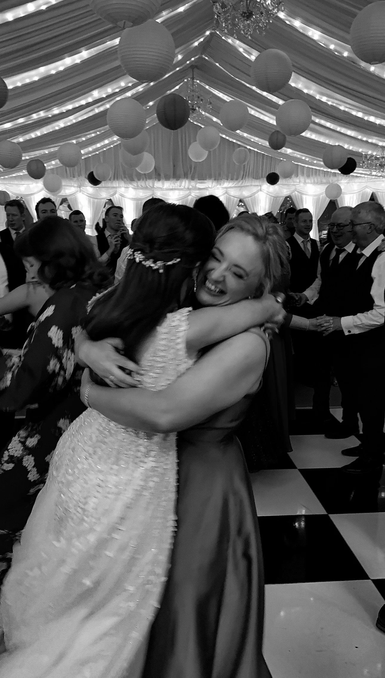 Two women hugging at a wedding reception with guests dancing in the background, decorated with hanging lanterns and string lights. Wedding Content Creator Ireland in Clonabreany House Wedding Venue
