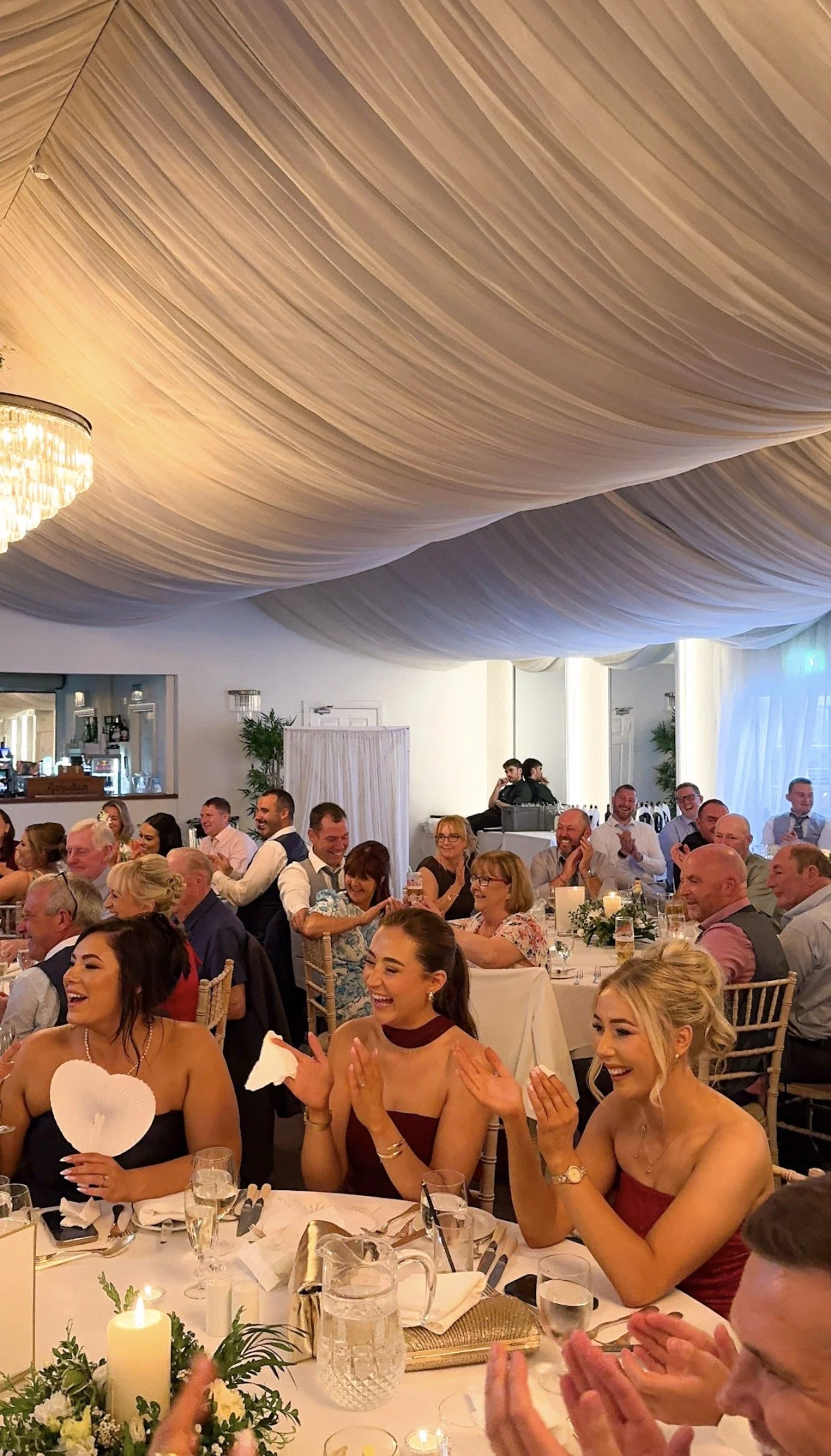 Guests at a wedding reception sitting at tables, laughing and clapping, decorated with flowers and candles, with a ceiling draped with fabric in Rathsallagh House, Wicklow Irish wedding venue.