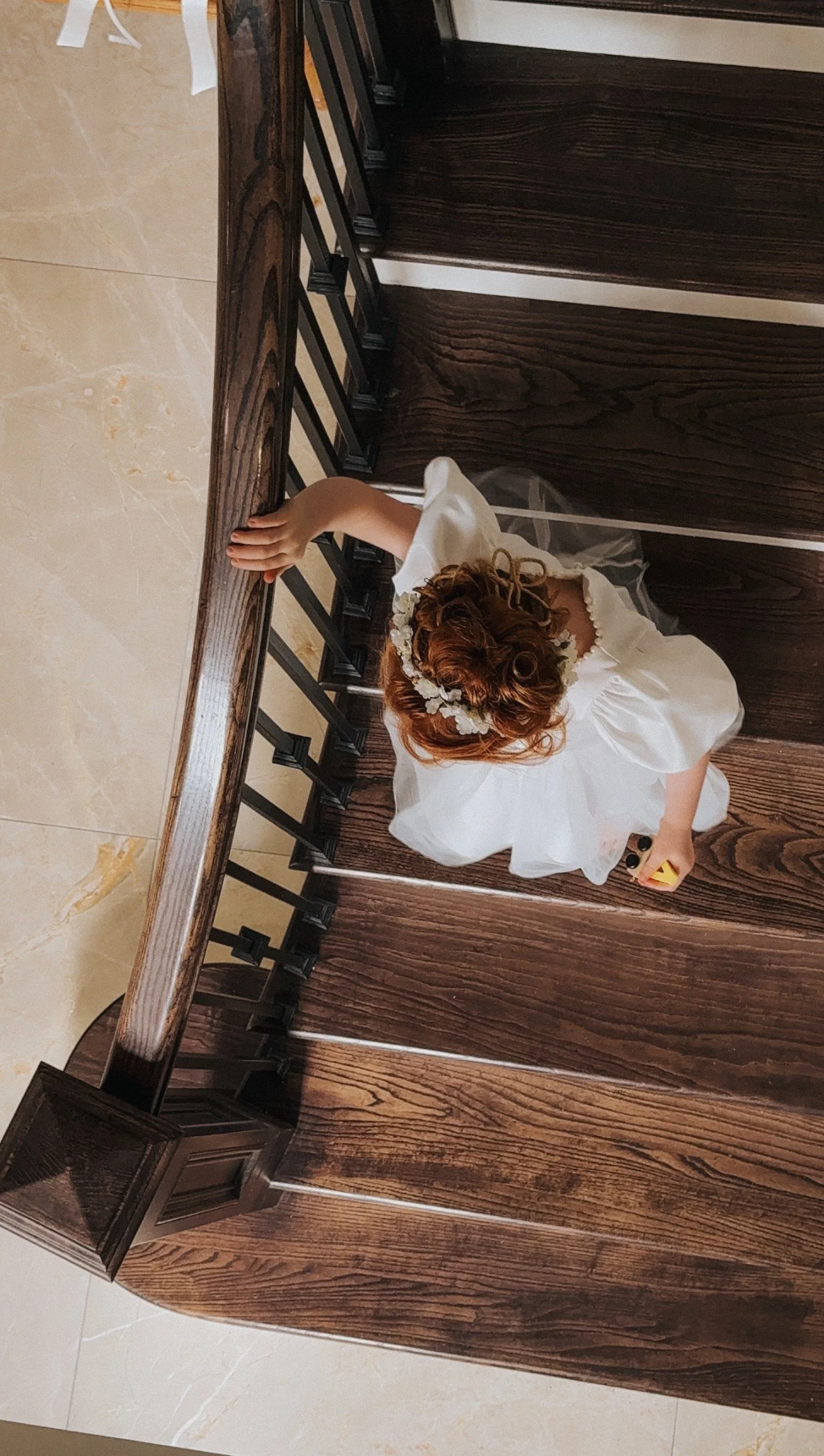 A young flower girl with red hair, wearing a white dress and a floral headband, is standing behind a wooden railing, viewed from above. In Co. Sligo, Wedding Content Creator Ireland 