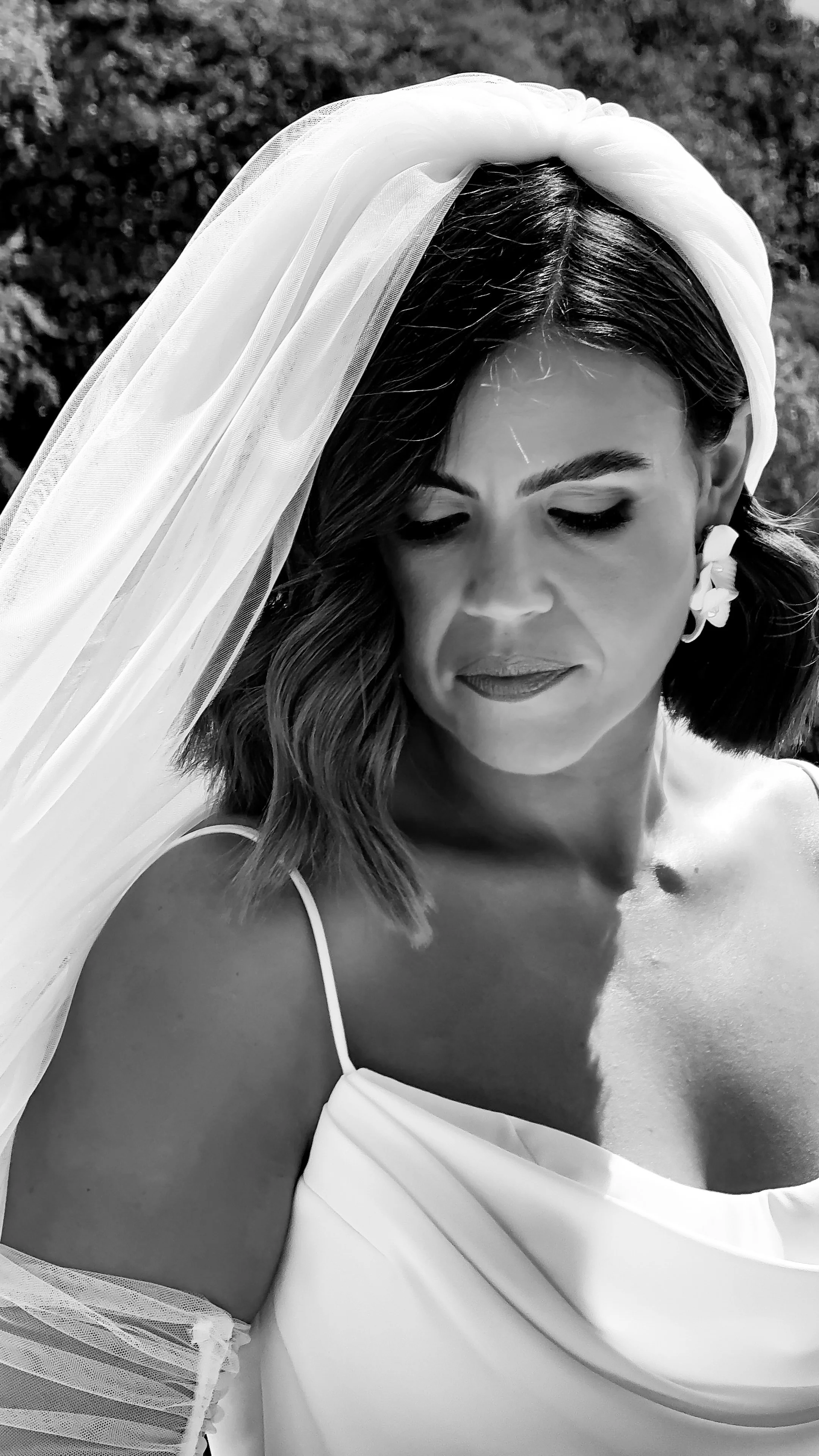 A black and white close-up photo of a woman in a wedding dress with a veil, outdoors, looking down, with a background of trees in Armagh.