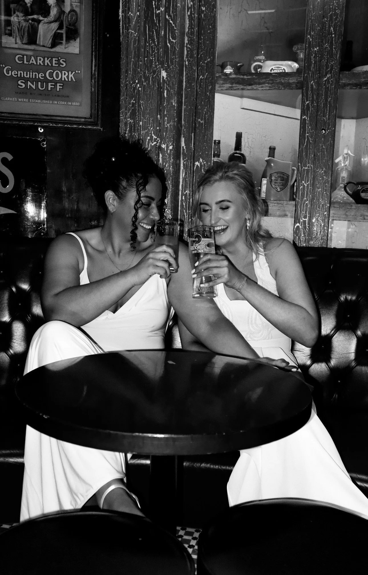Two brides in white dresses sharing a laugh and clinking glasses in a bar, with a wooden wall and framed pictures in the background at their Dublin City Wedding. Dublin Wedding Content Creator