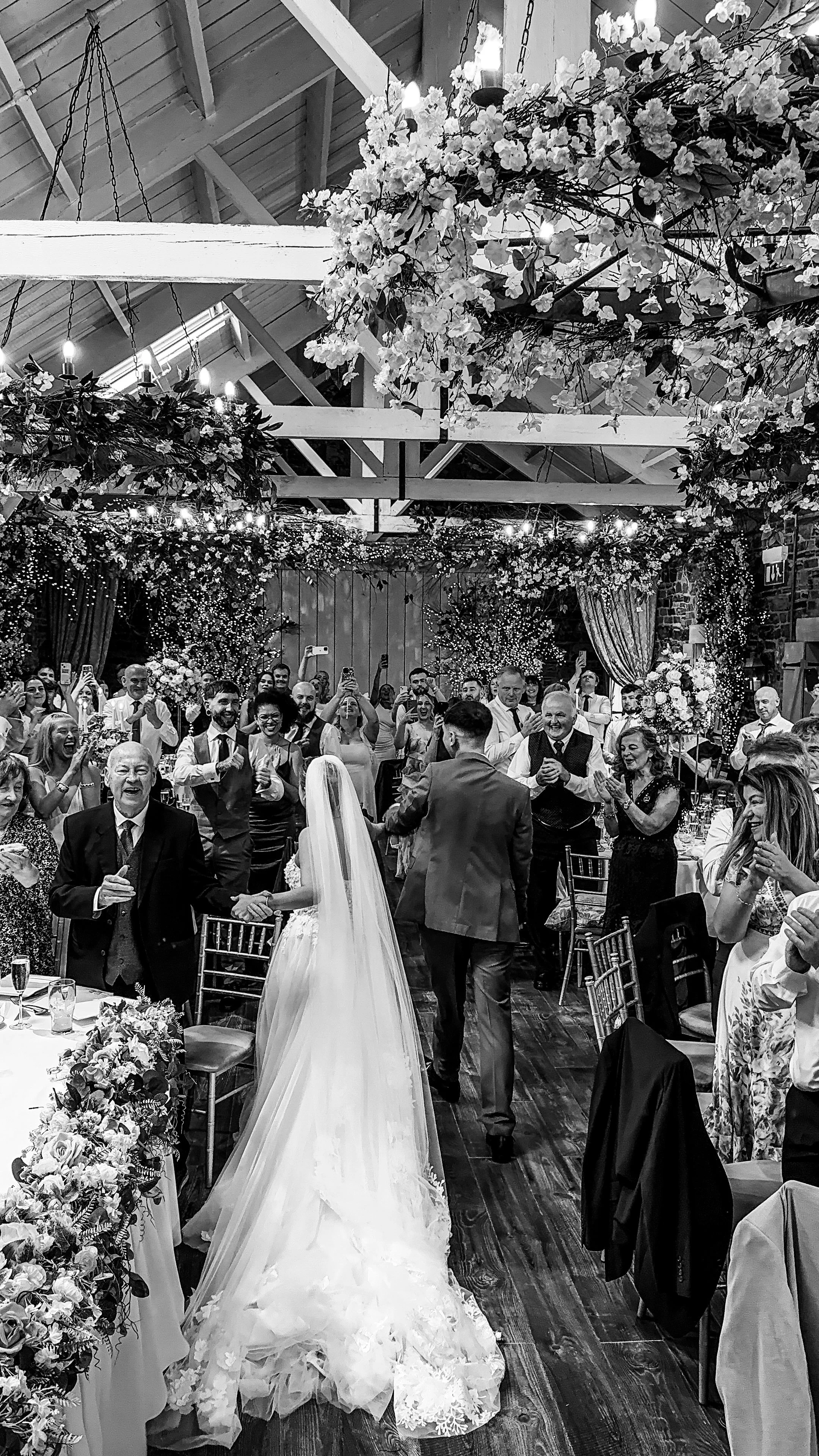 A joyful wedding reception with the bride and groom dancing, surrounded by guests in a decorated venue with floral arrangements and hanging chandeliers.