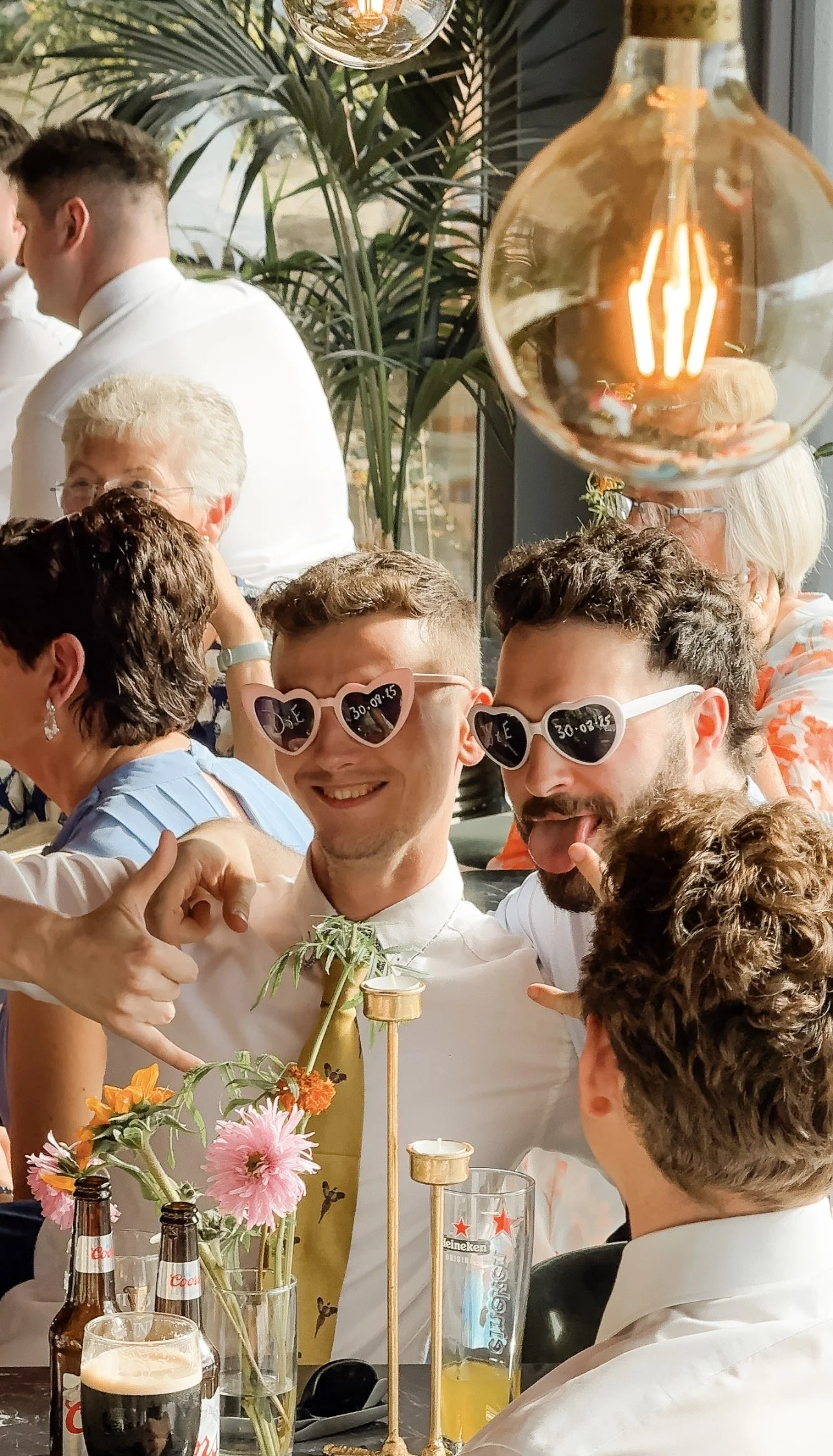 Group of people celebrating, wearing sunglasses, with flowers and drinks on the table, indoors with greenery and hanging light bulb decorations in Mountain View Kilkenny.