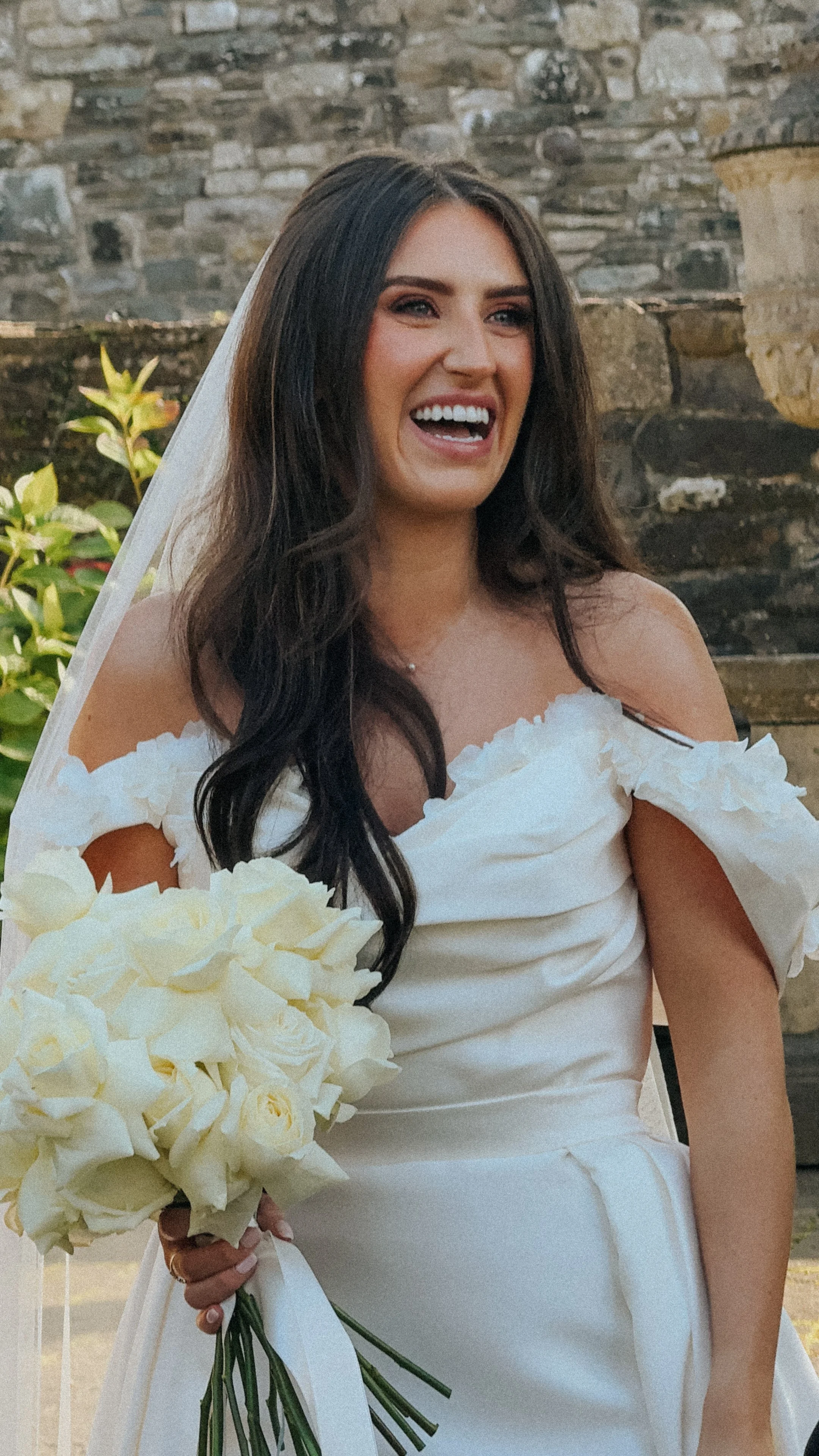 A bride with long dark hair, wearing a white wedding dress with off-the-shoulder ruffled sleeves, holding a bouquet of white roses, standing outdoors in front of a stone wall in Tankardstown House. Meath Wedding Content Creator