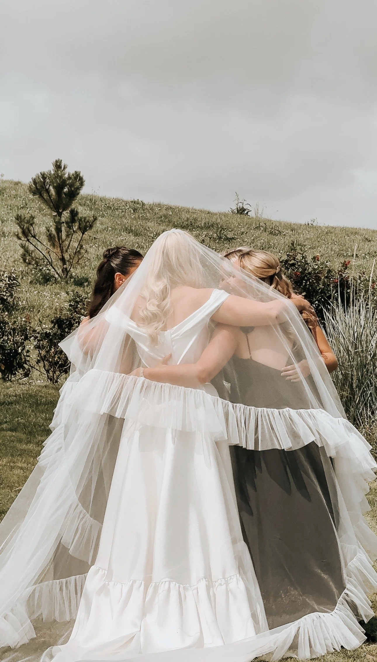 Three women, one is a bride in a white wedding gown, sharing an embrace under a sheer veil outdoors with a grassy hill and small trees in the background in Co. Cork. Wedding Content Creator Ireland 