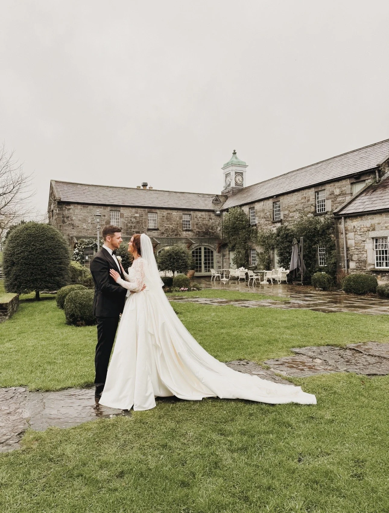 A bride and groom dancing outdoors on a rainy day at a historic stone building estate, with lush green grass and wet stone paths in Ballymagarvey Village, Co. Meath Wedding Venue. Wedding Content Creator Ireland 