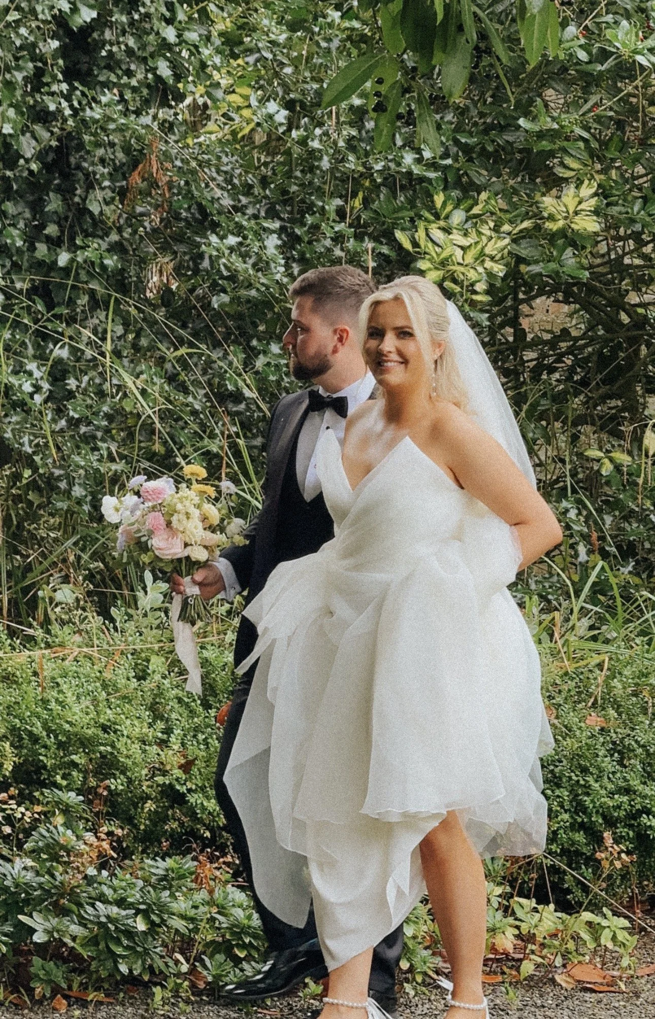 A bride and groom posing outdoors in Rathsallagh House, Co. Wicklow, Wedding Venue. The groom holding a bouquet of pink and cream flowers, both dressed in wedding attire, smiling and enjoying the moment. Wedding Content Creation Ireland
