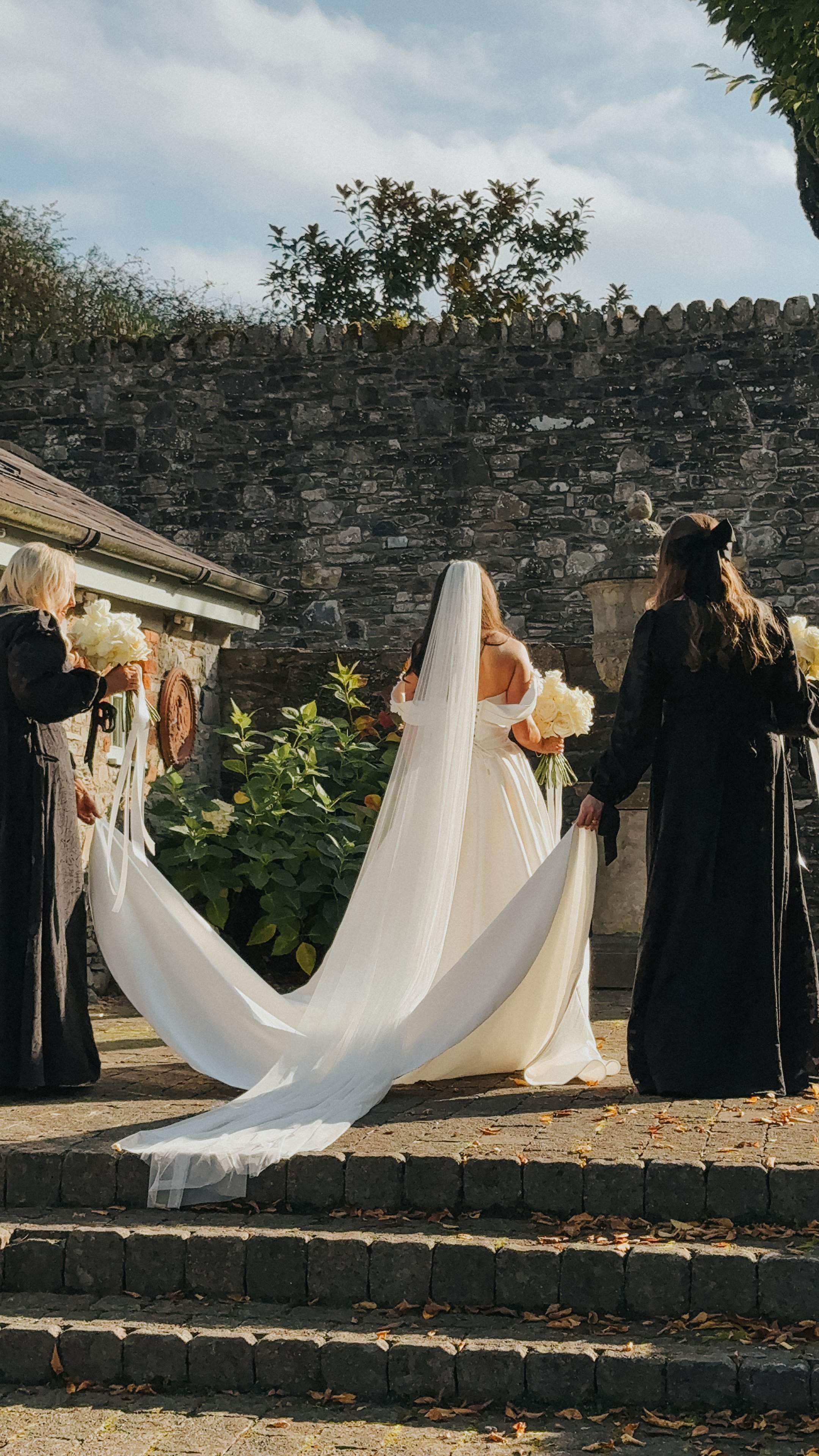 An Irish bride in a white wedding dress and veil holding a bouquet, standing on outdoor stone steps, with three women, two in black dresses and one in dark dress, surrounding her, holding flowers. Wedding Content 