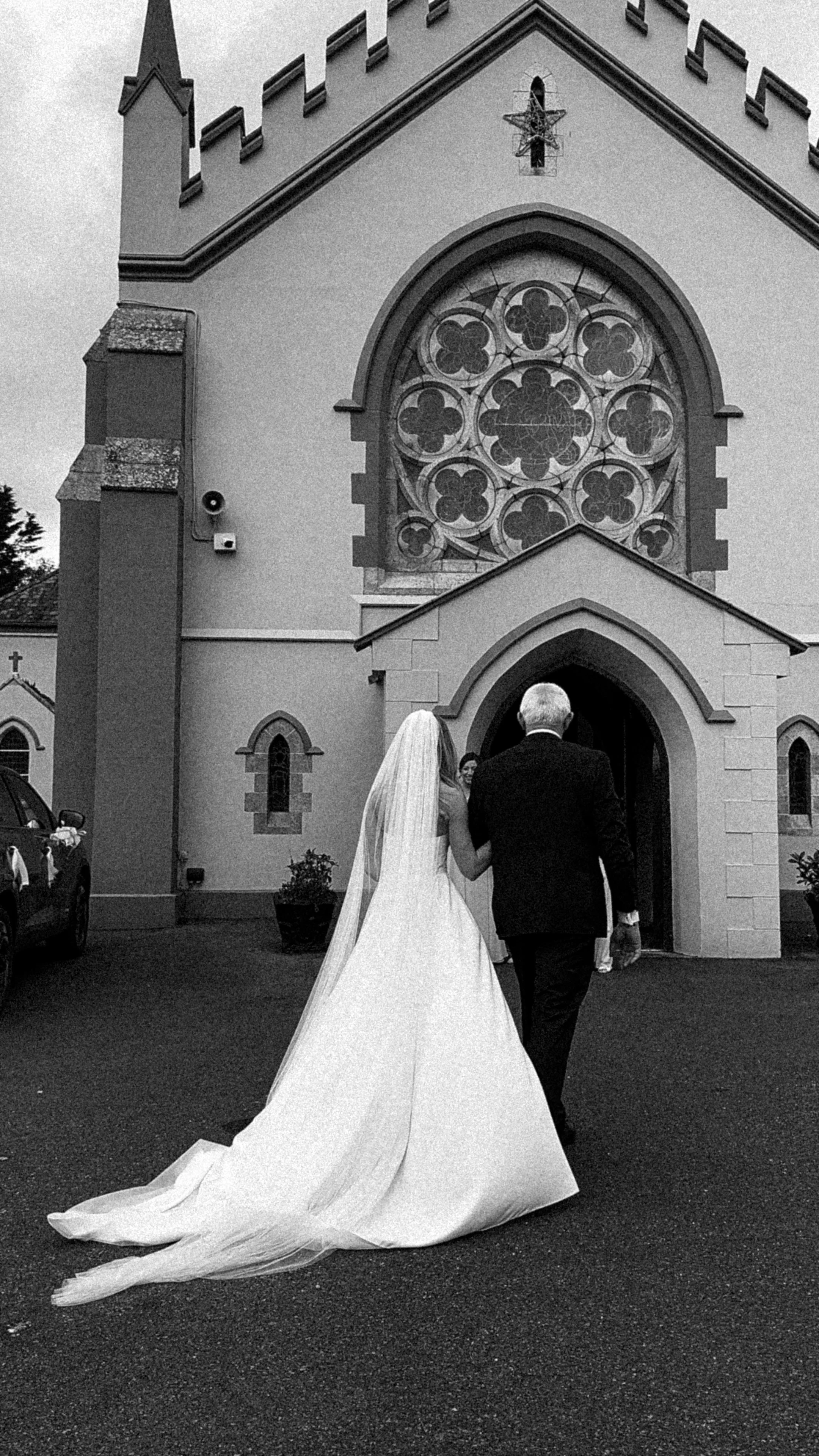 A bride and an older man, possibly her father, walking toward a church entrance on her wedding day. The church has a large stained glass window and Gothic architectural details in County Sligo. Wedding Content Ireland.