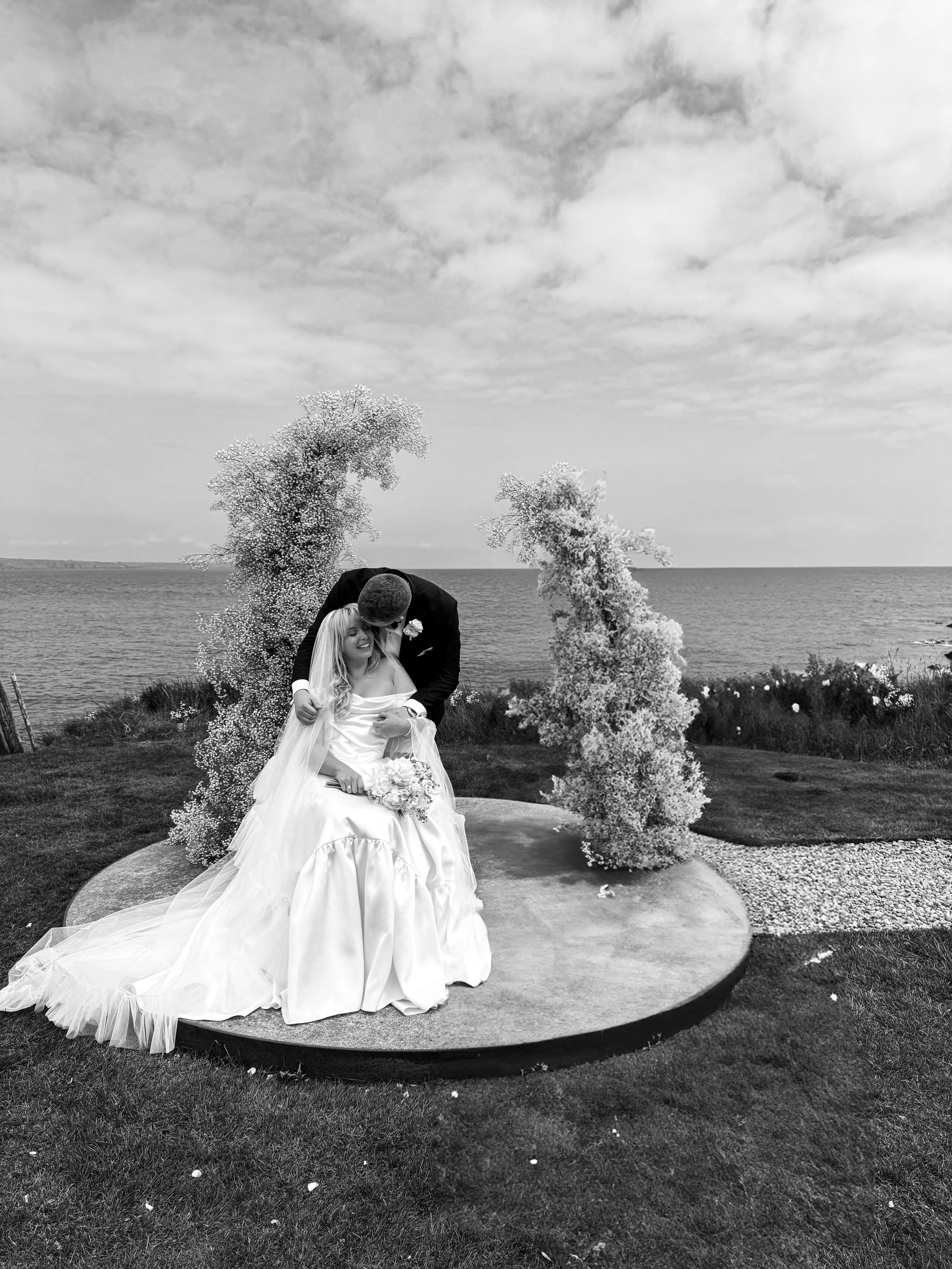 A bride and groom on a wedding day in Cork, standing on a circular platform outdoors near the ocean, with trees and a cloudy sky in the background. The bride is sitting, holding a bouquet, and smiling while the groom leans over her, holding her face.