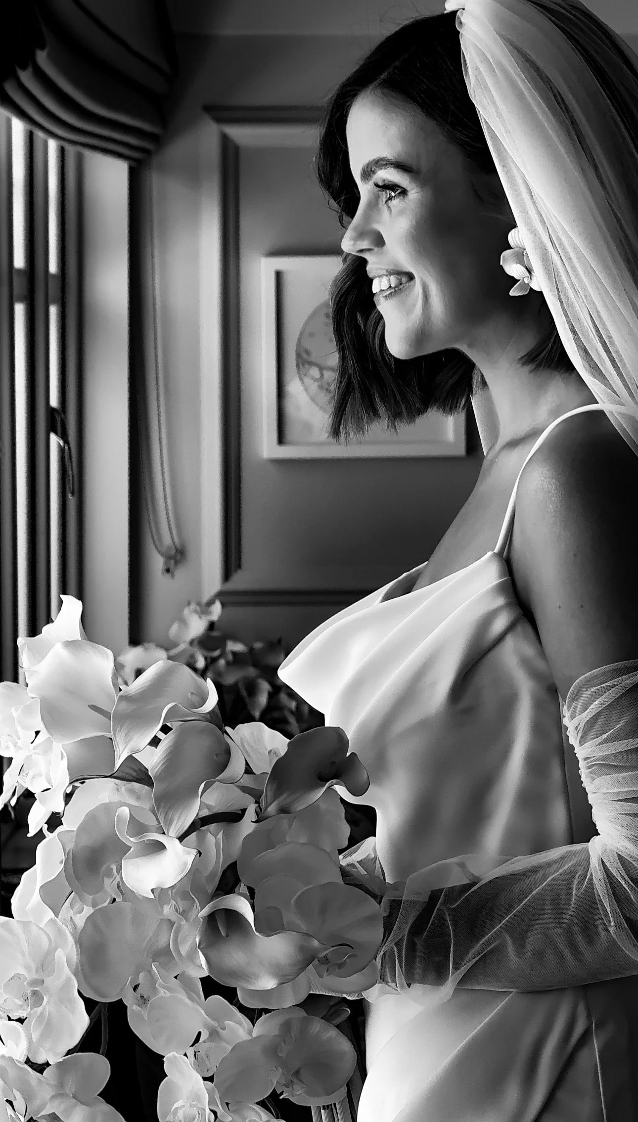 Black and white photo of a woman with short hair, smiling, wearing a wedding dress and veil, holding a bouquet of flowers, looking out a window in The Station house wedding venue in County Meath