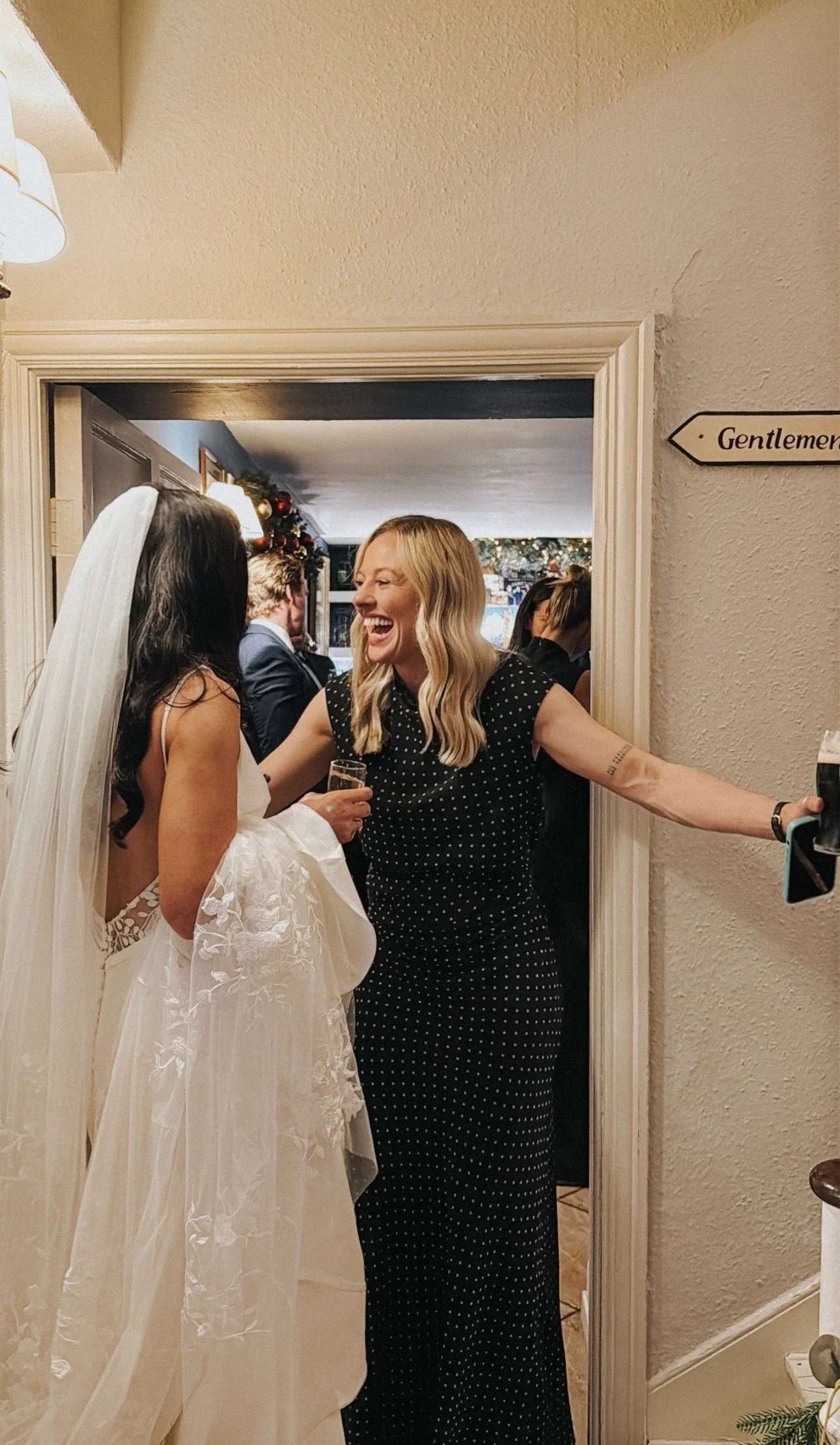 A woman in a wedding dress and veil talking to a woman in a black dress with white polka dots, both smiling and laughing at a wedding reception in Rathsallagh House, Wicklow.