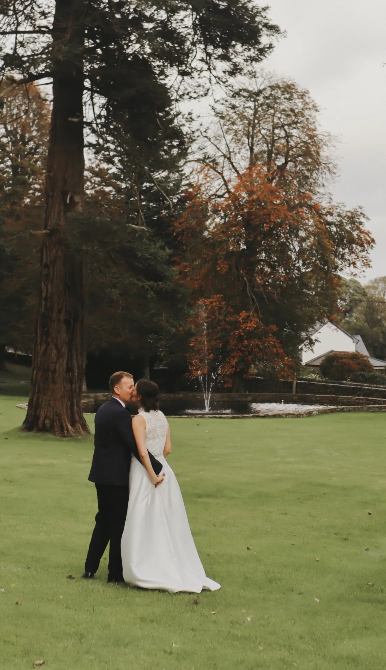 A newlywed couple kissing on a lawn in a park with large trees and a fountain in the background. In Drumhalla House, Wedding Venue in Donegal. Irish Wedding Content Creator - Grey Acre Studios