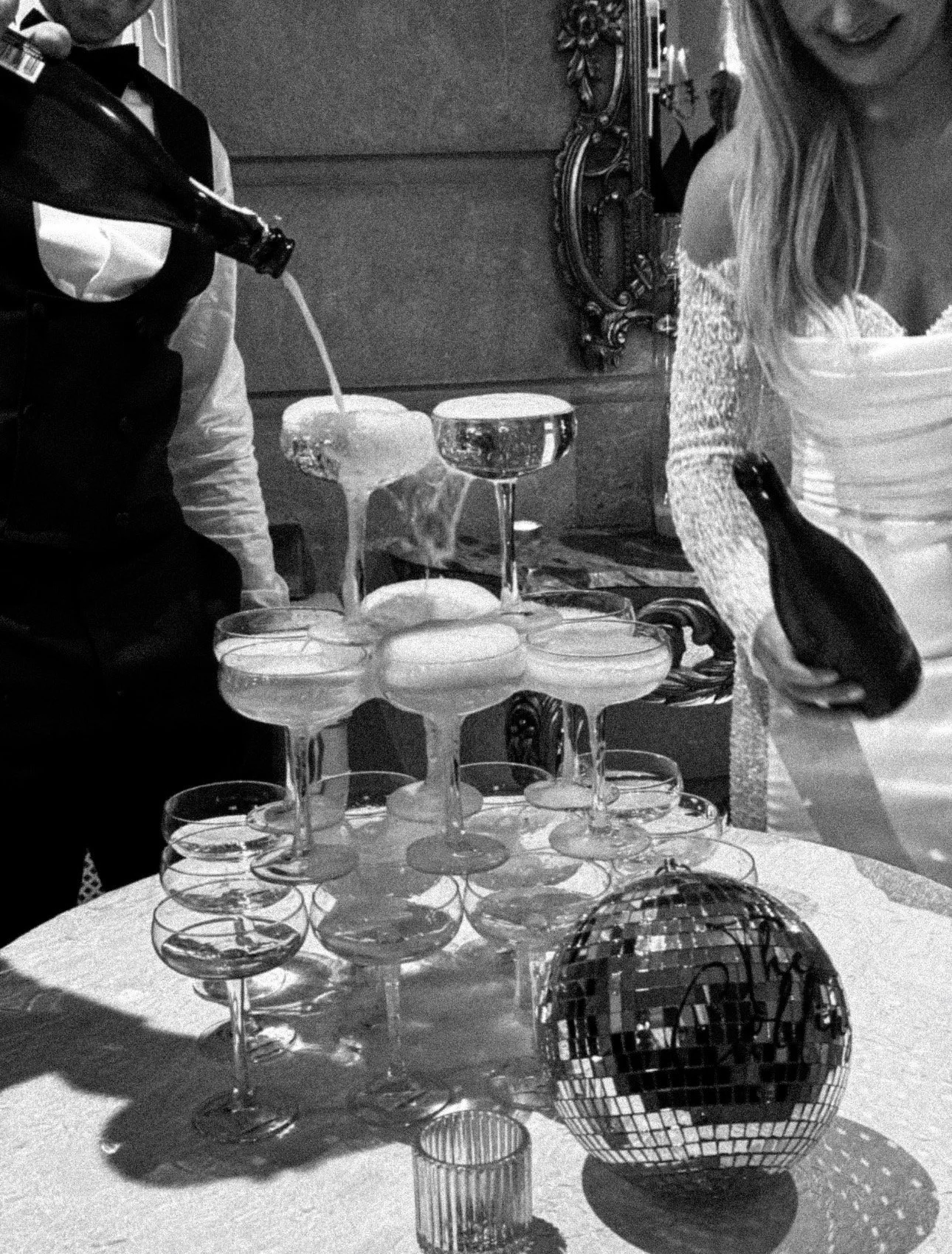 A black and white photo of a champagne tower made of coupe glasses on a table, with a woman holding a bottle of champagne and a disco ball nearby.