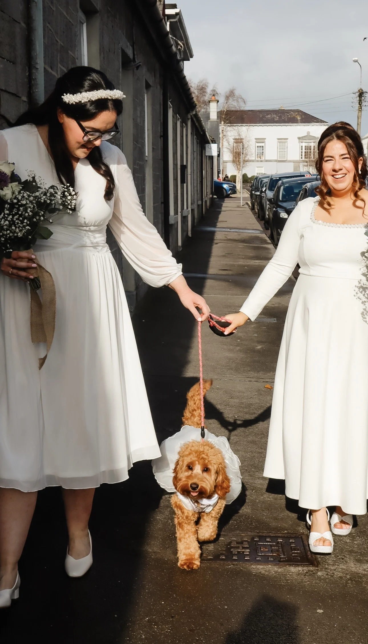 Two women in white dresses holding a small dog dressed in a white outfit on a city sidewalk, smiling and holding hands in Co. Kerry. Wedding Content Creator Ireland 