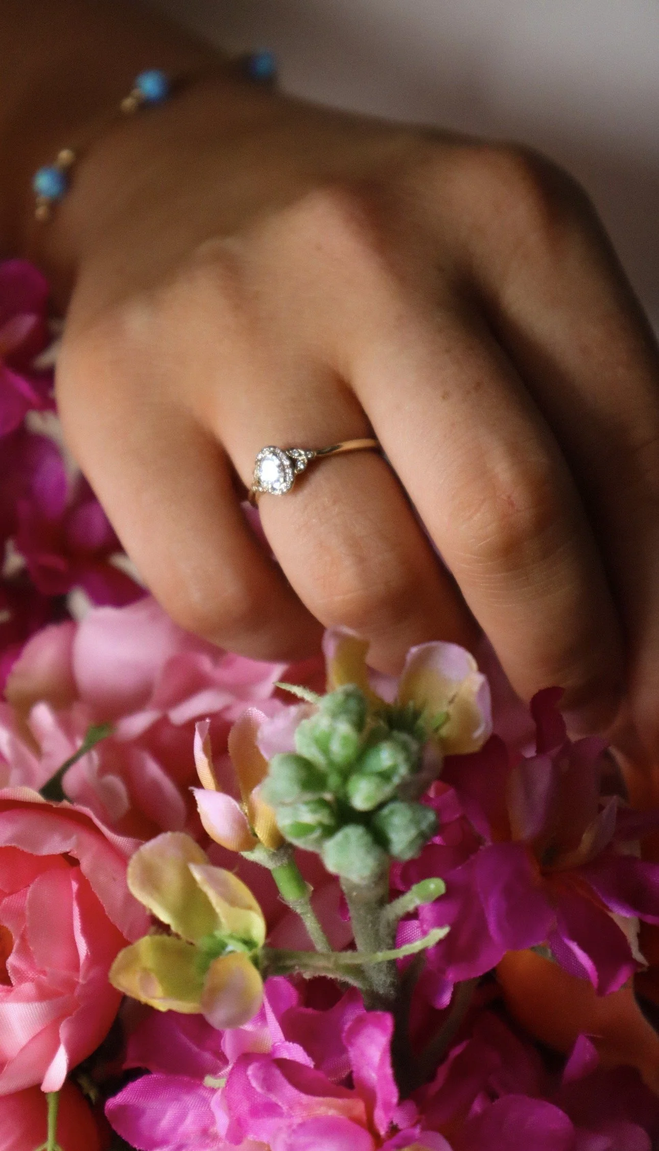 Close-up of a hand with a diamond ring on the ring finger, surrounded by pink and green flowers.