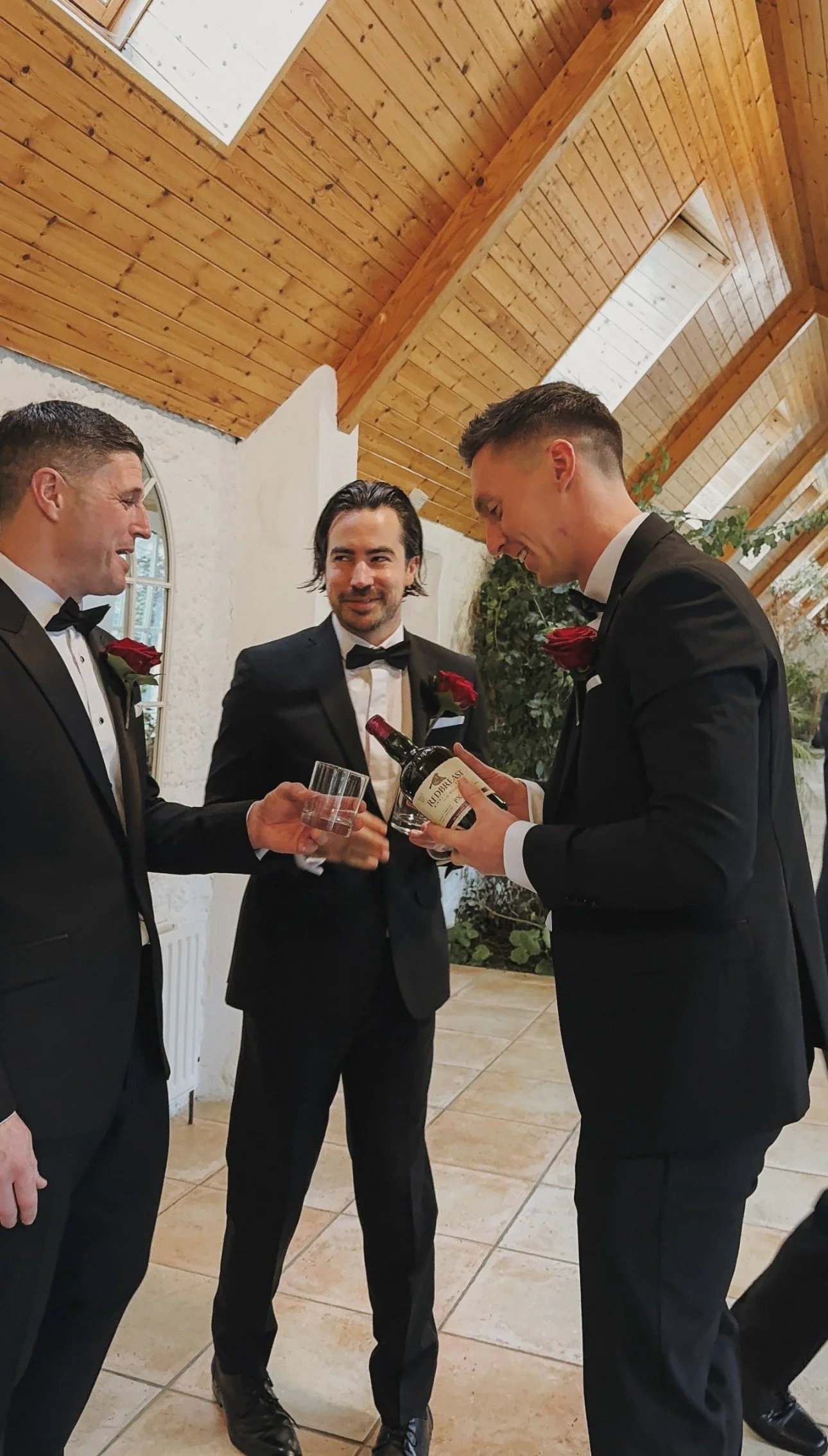 Three men in tuxedos with red roses on their lapels are gathered in a room with a wooden vaulted ceiling. One man is pouring wine into a glass held by another, while the third man watches with a smile. Wedding Content Creator Ireland 