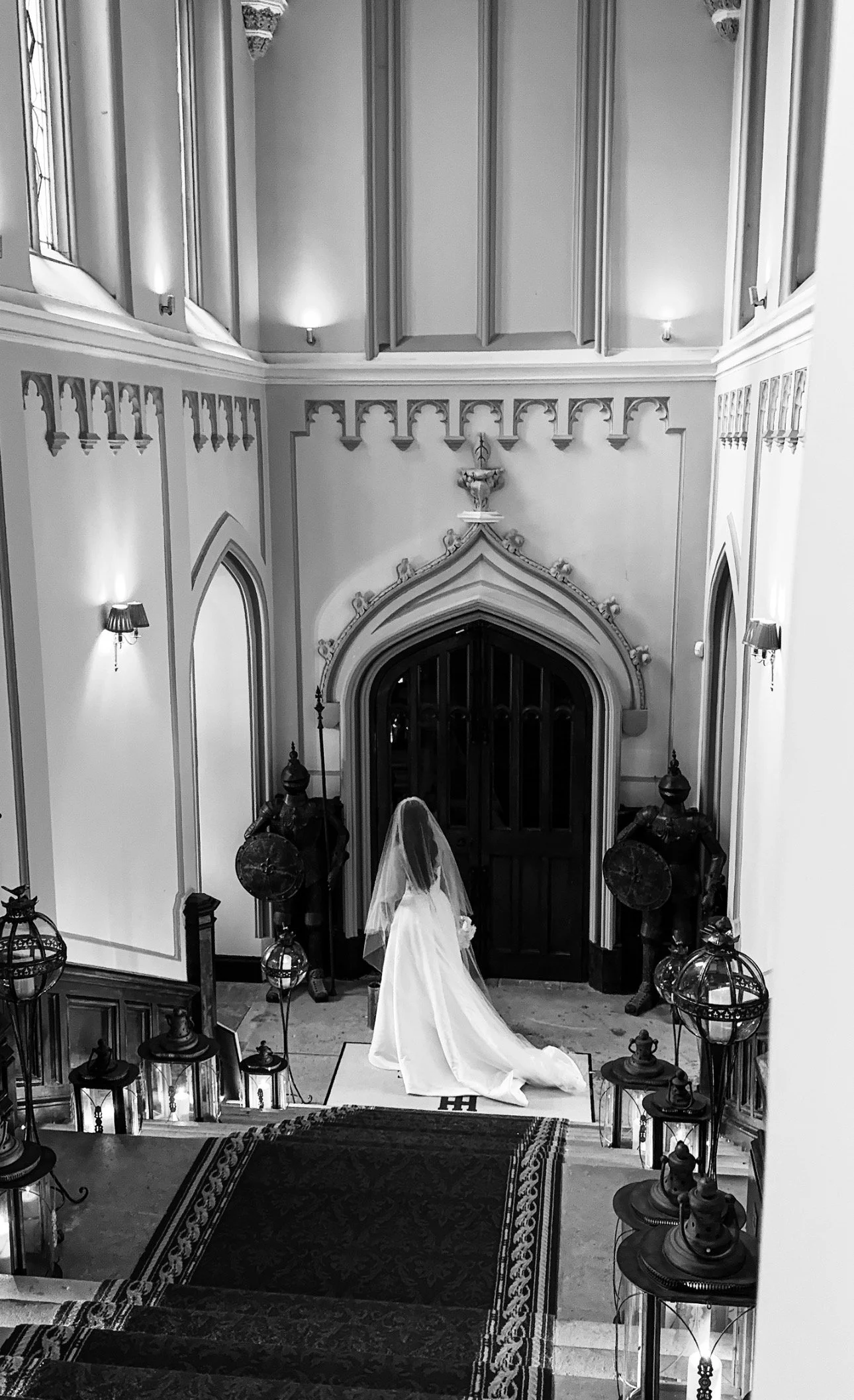 A bride in a flowing wedding gown and veil stands at an ornate wooden door inside a decorated gothic-style chapel, surrounded by lanterns and knight statues in Markree Castle, Sligo