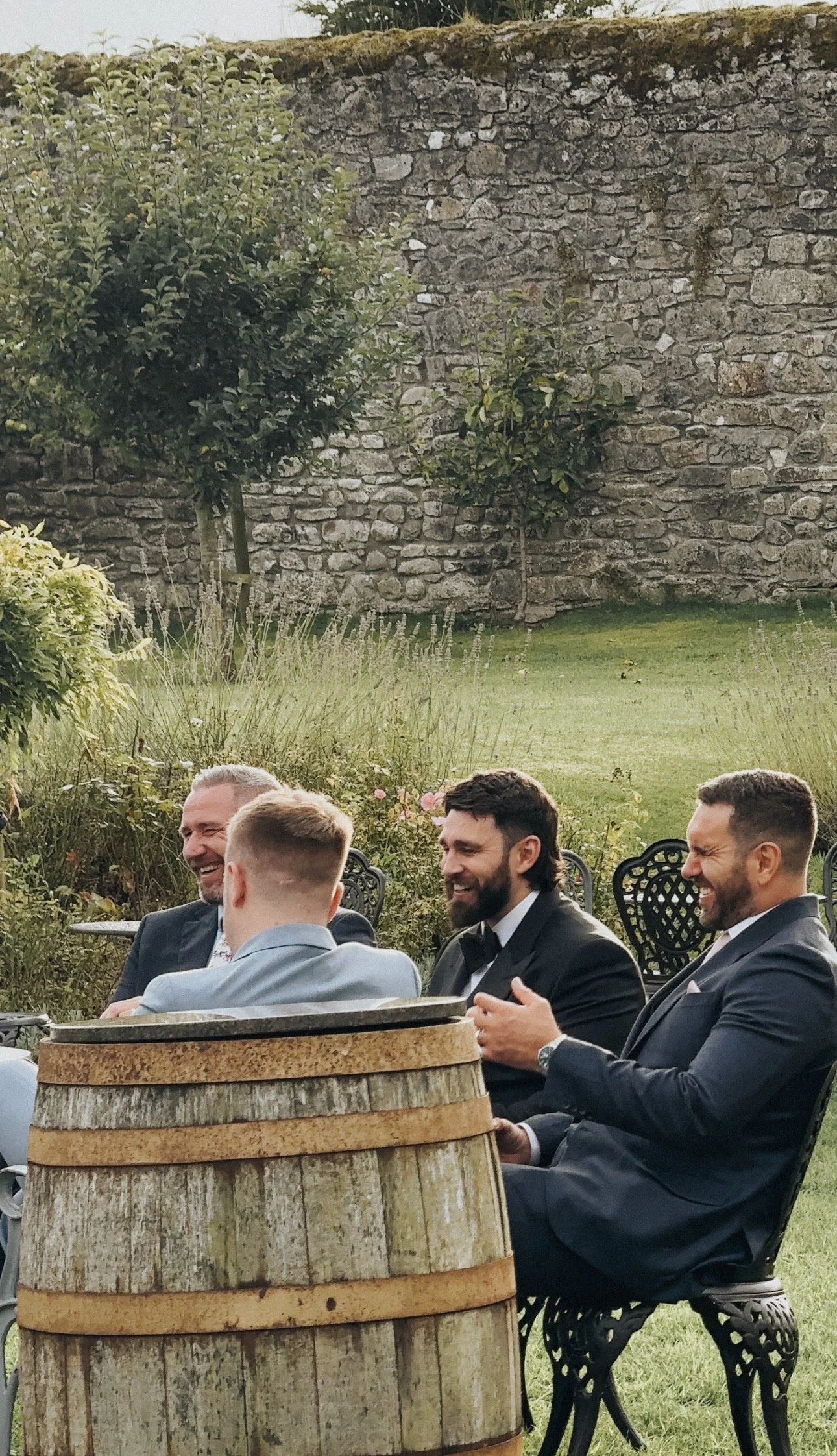 Four men in suits sitting and laughing outdoors during a garden party or wedding, with a stone wall, green grass, plants, and trees in the background in Ballymagarvey Wedding Venue, Meath. Wedding Content Creator Ireland 