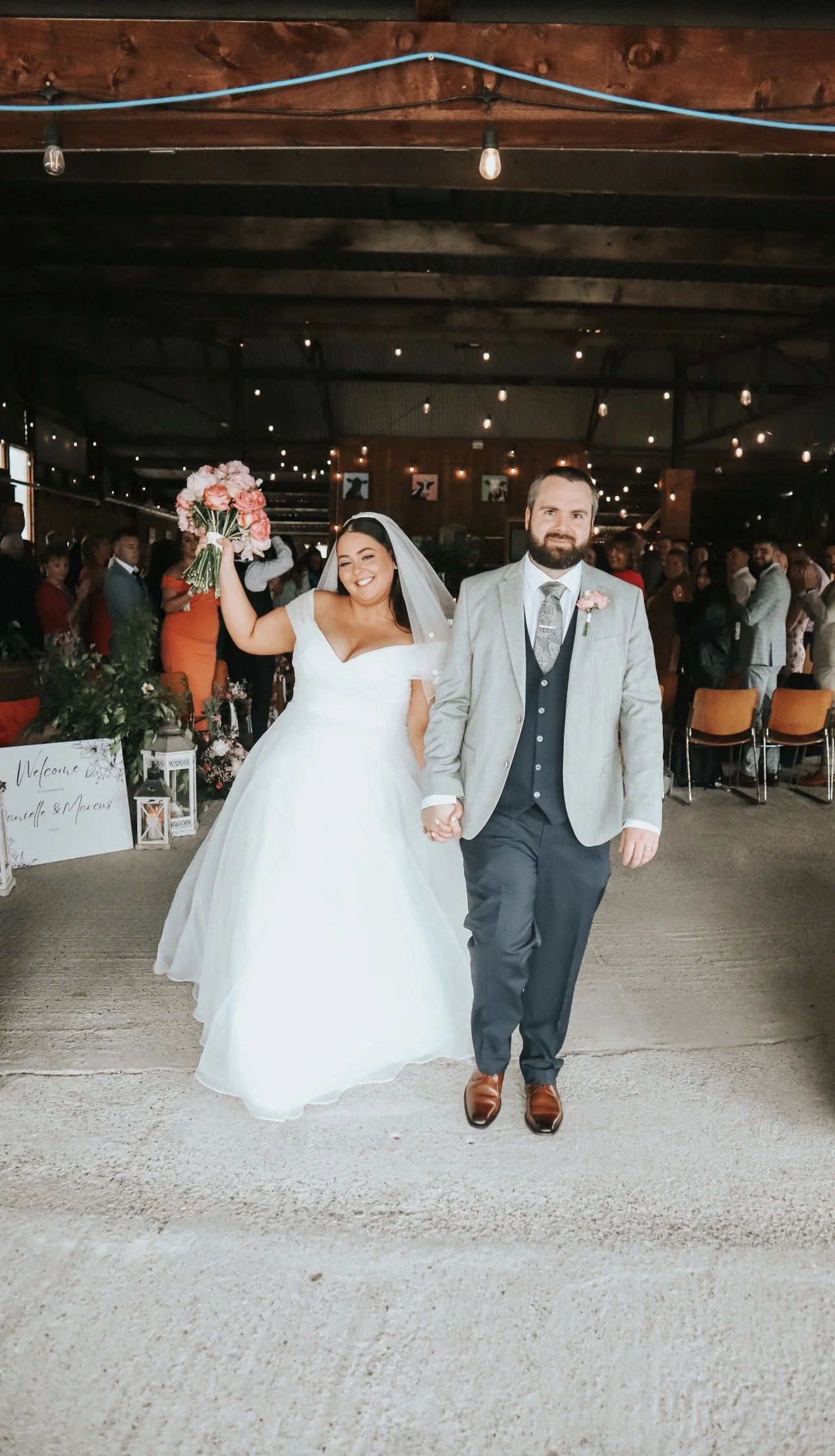 Bride and groom walking hand in hand at their wedding reception in Mountain View Kilkenny with guests in the background. The bride is holding a bouquet of pink flowers. The groom is dressed in a light gray suit with a boutonniere.
