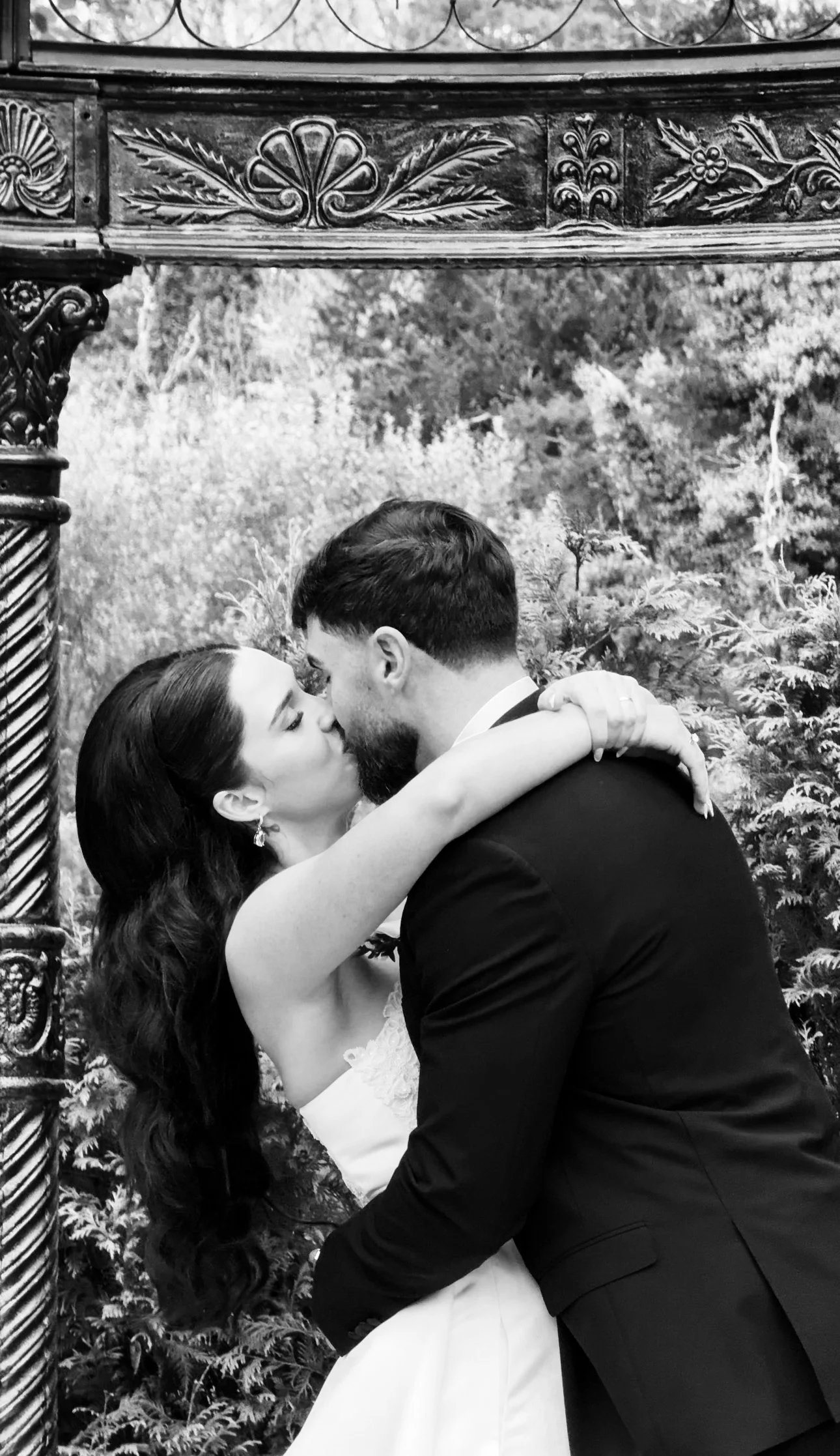A black-and-white photo of a couple kissing, with the woman wearing a white dress and the man wearing a dark suit, outdoors with greenery in the background. Captured in Markree Castle, Sligo by Wedding Content Creator Grey Acre Studios 