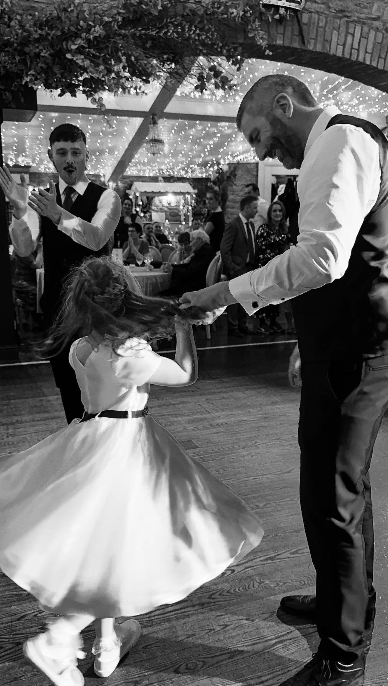 A young girl in a white dress spinning on the dance floor while holding hands with a man in a tuxedo. A man in a vest and tie looks on with surprise, and guests seated at tables are watching in the background. Wedding venue is Darver Castle Co. Louth