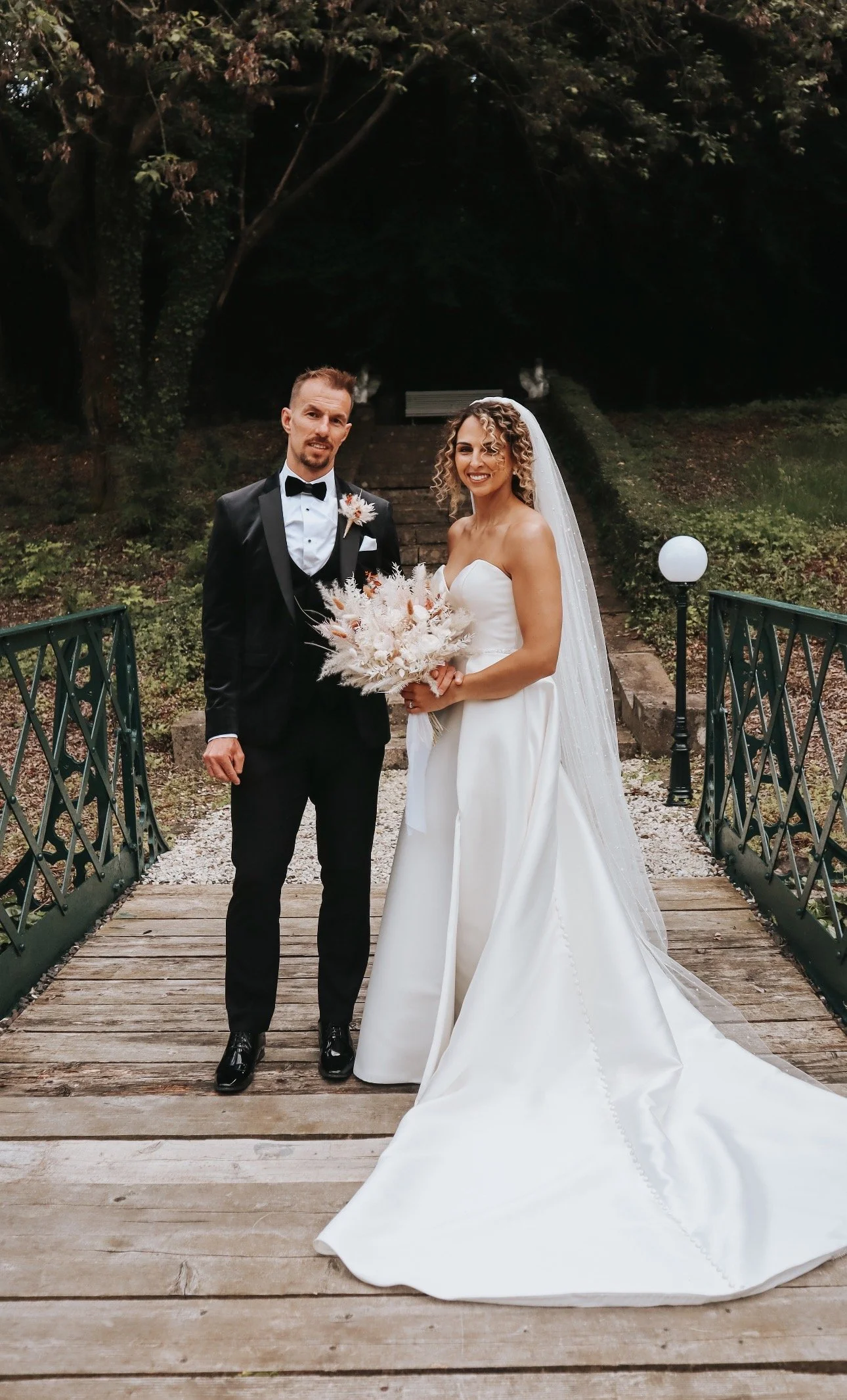 Bride and groom in wedding attire standing on a wooden bridge outdoors with greenery, trees, and stairs in the background in Boyne Hill House, Meath. Irish Wedding venue, Meath Wedding Content Creator Ireland 