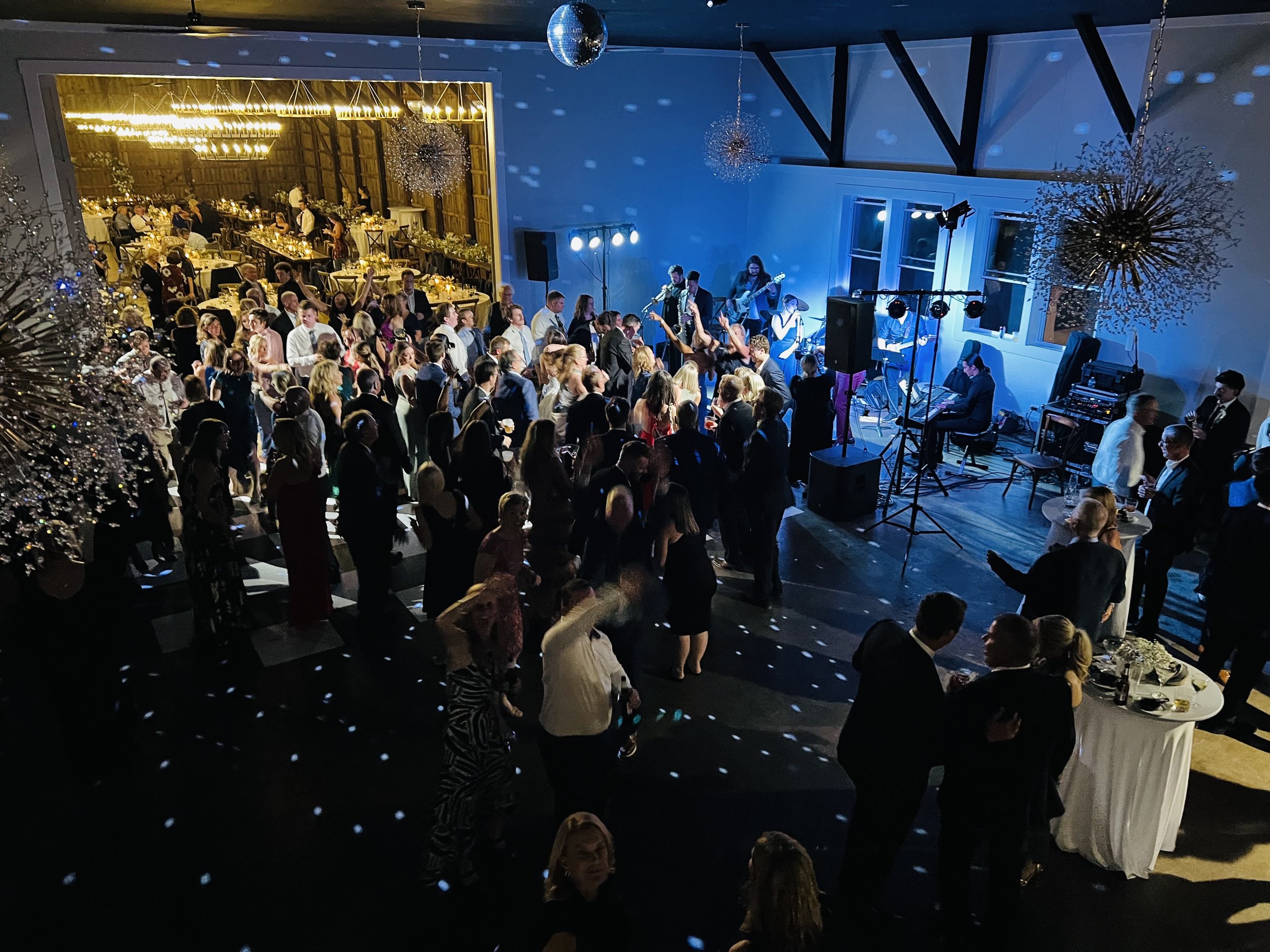People dancing and socializing at a lively indoor party or wedding reception with band playing on stage. Guests are wearing formal attire, and decorations include hanging ornaments and table settings.