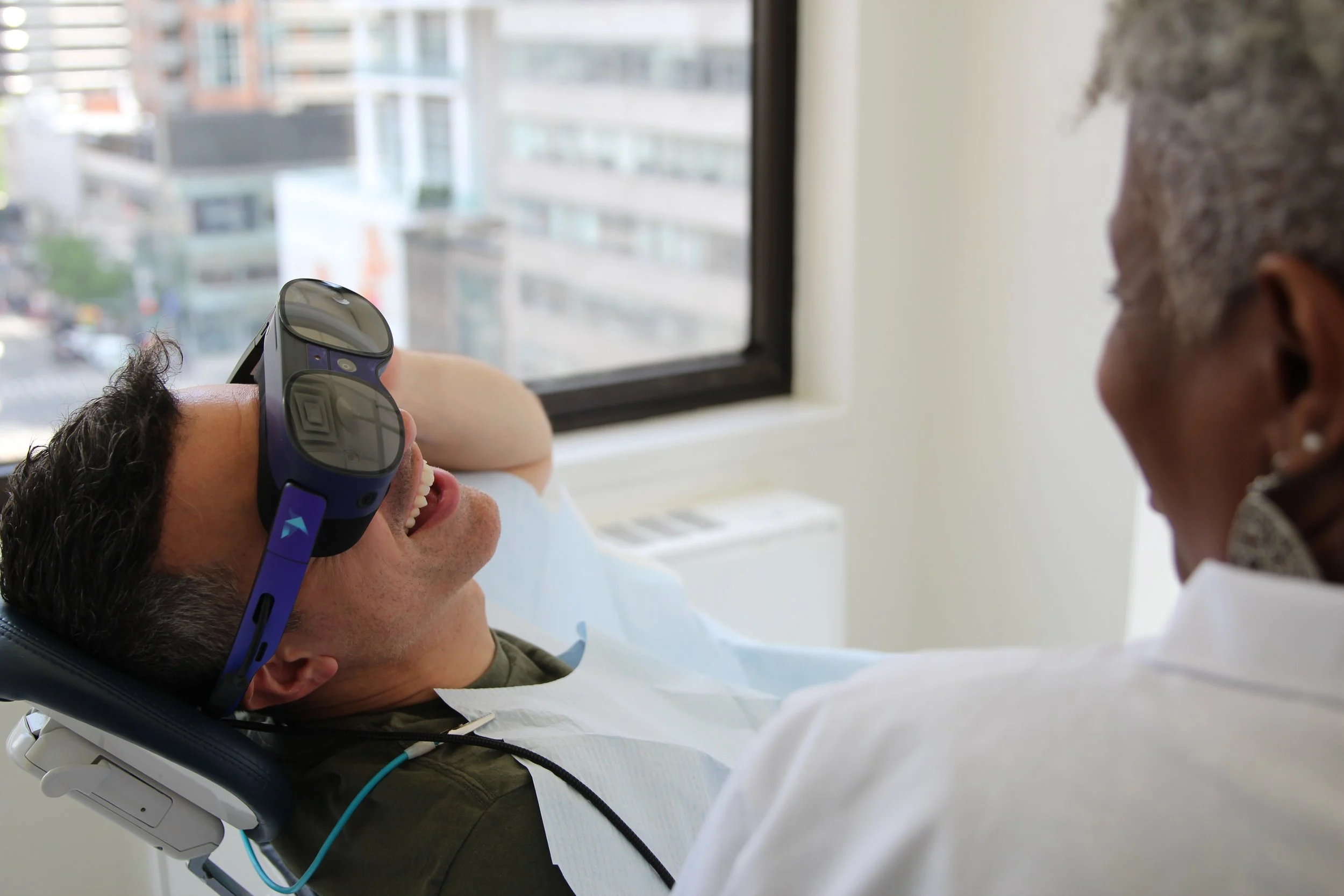 A man lying on a medical chair wearing eye protection glasses, talking and smiling, with a healthcare worker beside him in a clinical setting near a window.