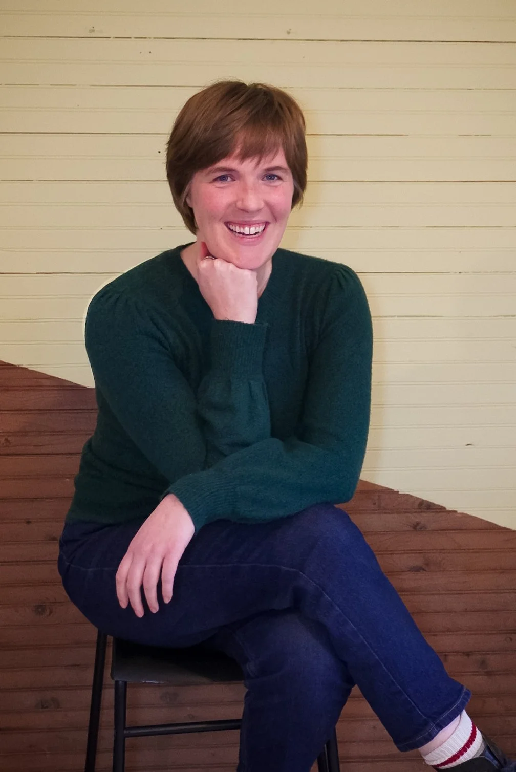 A woman with short brown hair, wearing a dark green sweater and dark jeans, sitting on a wooden chair with one leg crossed over the other, smiling and resting her chin on her hand against a light-colored wooden wall.