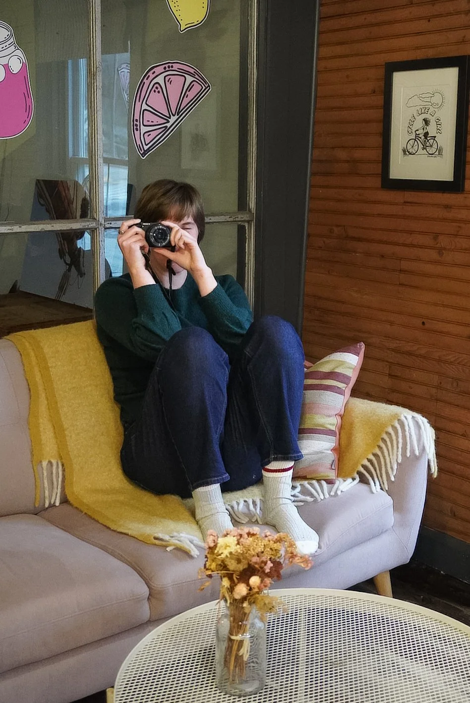 A person sitting on a beige couch, holding a camera up to their eye and taking a photo. They are wearing a dark green sweater, jeans, and striped socks. There's a yellow blanket and a striped pillow behind them, with a small flower arrangement on a white round table in front. The room has wood-paneled walls and decorated with colorful fruit illustrations on a glass partition.