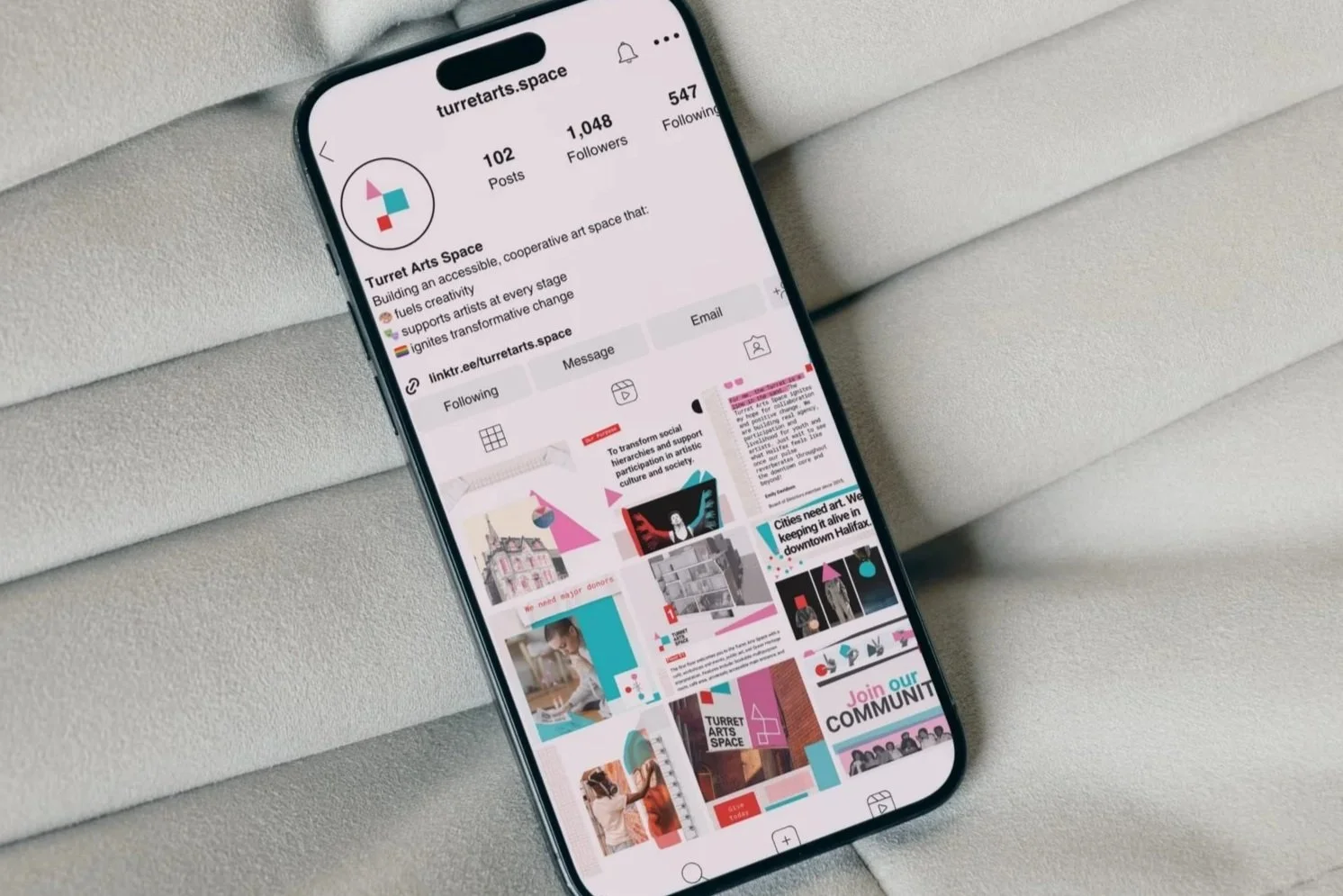 An iPhone displaying the Instagram profile of Turret Arts Space with 102 posts, 1,048 followers, and 547 following, resting on a light-colored textured fabric surface.
