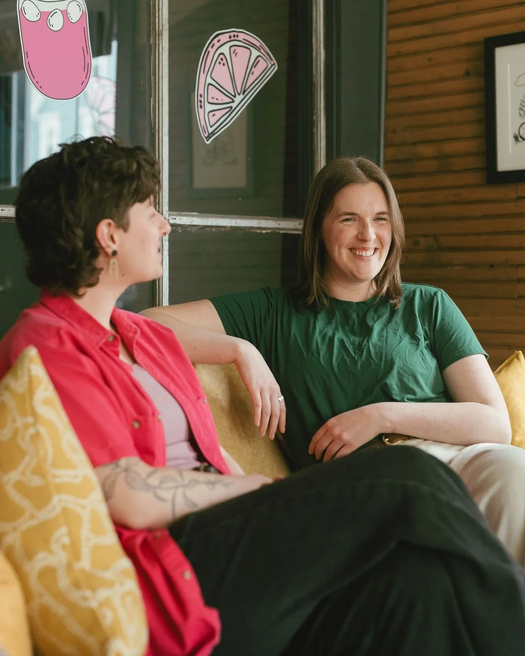 Meghan and Emily sitting on a yellow couch, smiling and engaging in conversation, with playful pink and lemon slice window stickers behind them.