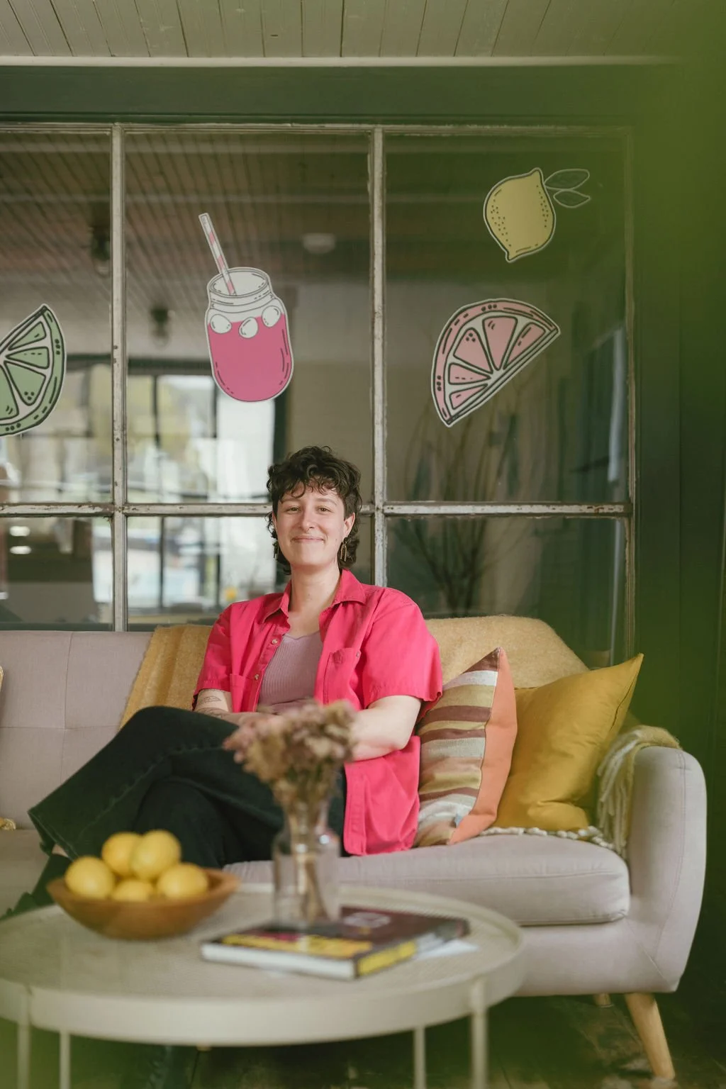 Meghan, a brand and website designer, is sitting on a couch in her office. There are illustrated citrus fruit stickers on a window behind her