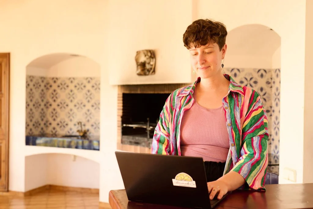 Meghan, a woman with short curly hair, wearing a colorful striped shirt and a pink tank top, smiling while working on a black laptop in a cozy living room.