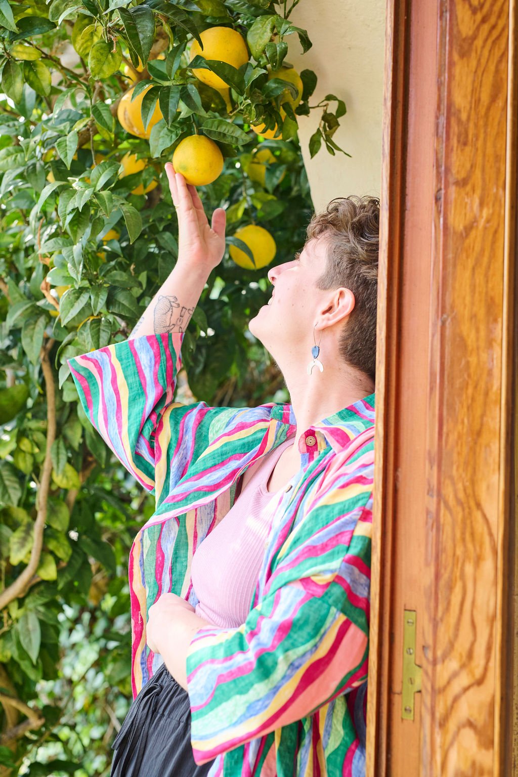 Meghan, a women with short hair and colorful striped shirt reaching up to pick a lemon from a tree in a garden.