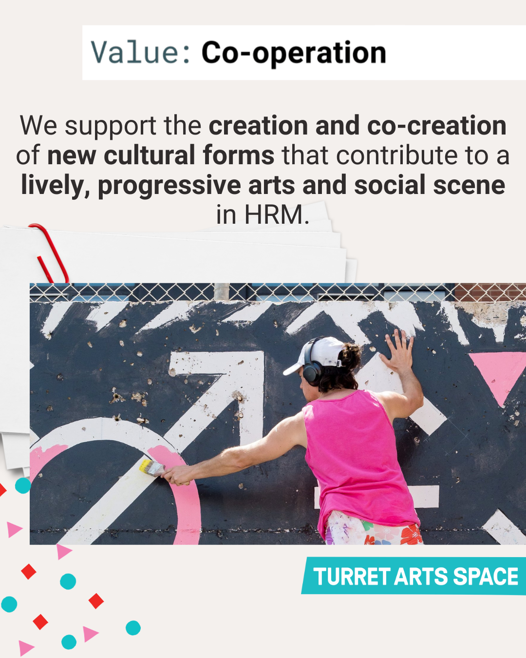 A person painting a mural on a black wall with pink and white designs, wearing a pink sleeveless shirt, colorful shorts, a white cap, and headphones, at an outdoor art space called 'Turret Arts Space'.