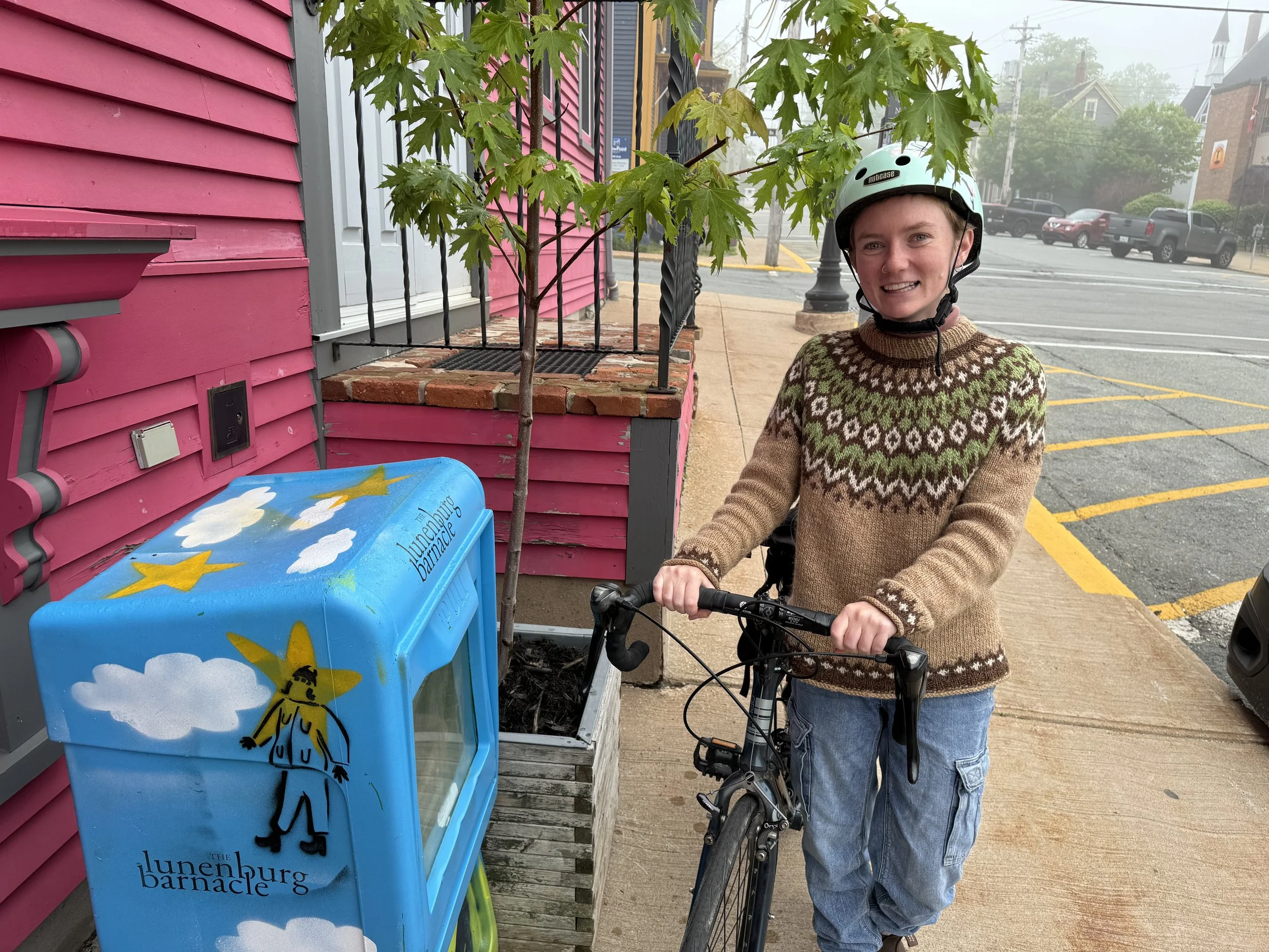 A smiling person in a biking helmet and sweater stands next to a bicycle under a tree on a foggy street, with a pink building in the background.