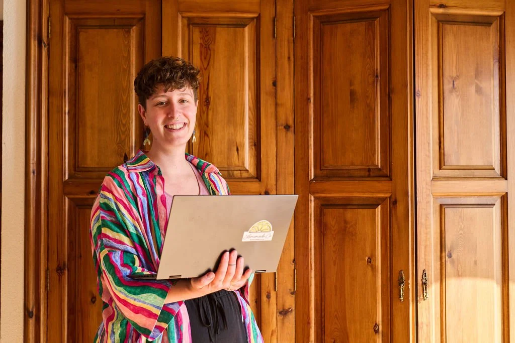 Meghan, a woman with short curly hair, wearing a colorful striped shirt, holding a laptop, and smiling in front of wooden closet doors.