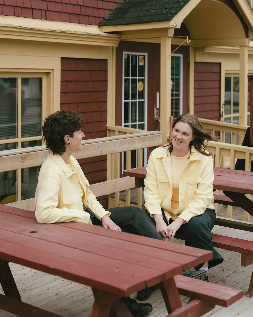 Meghan and Emily sitting on a wooden deck and engaging in conversation, both wearing light yellow jackets, with a red wooden building in the background.